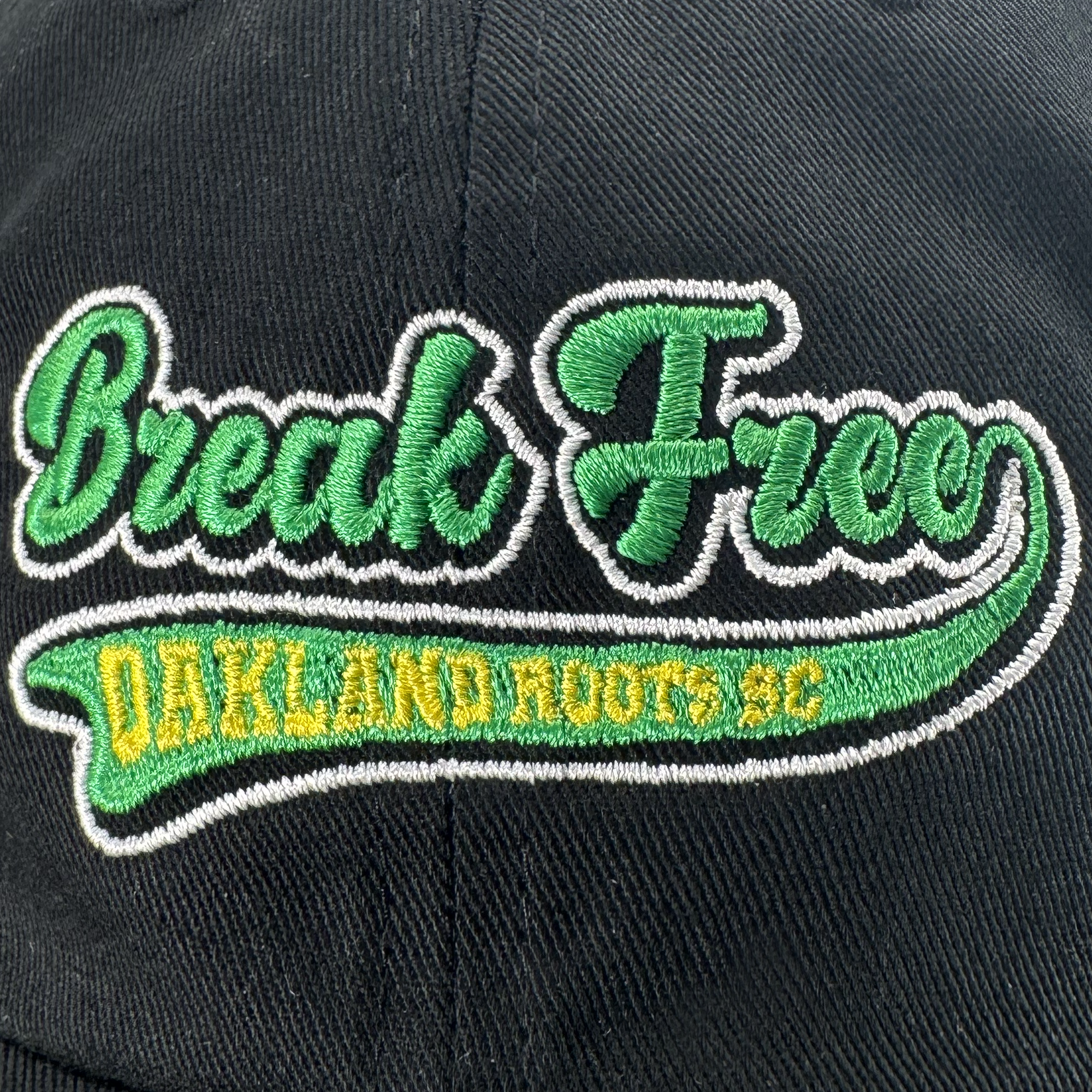 Close-up of the Oakland Roots SC X Break Free Hat by Oakland Roots SC, a black dad hat with bold green and white "Break Free" cursive embroidery and yellow-outlined "OAKLAND ROOTS SC" block letters. Detailed stitching emphasizes the design.