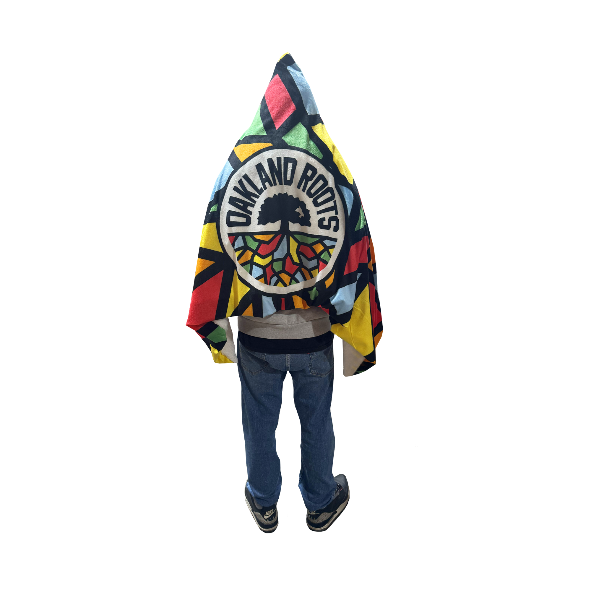 A person stands with their back turned, wrapped in the Oakland Roots SC Sherpa Blanket featuring a bold tree logo and geometric patterns. The cozy blanket, by Oakland Roots SC, pops against their hoodie and jeans on a transparent background.