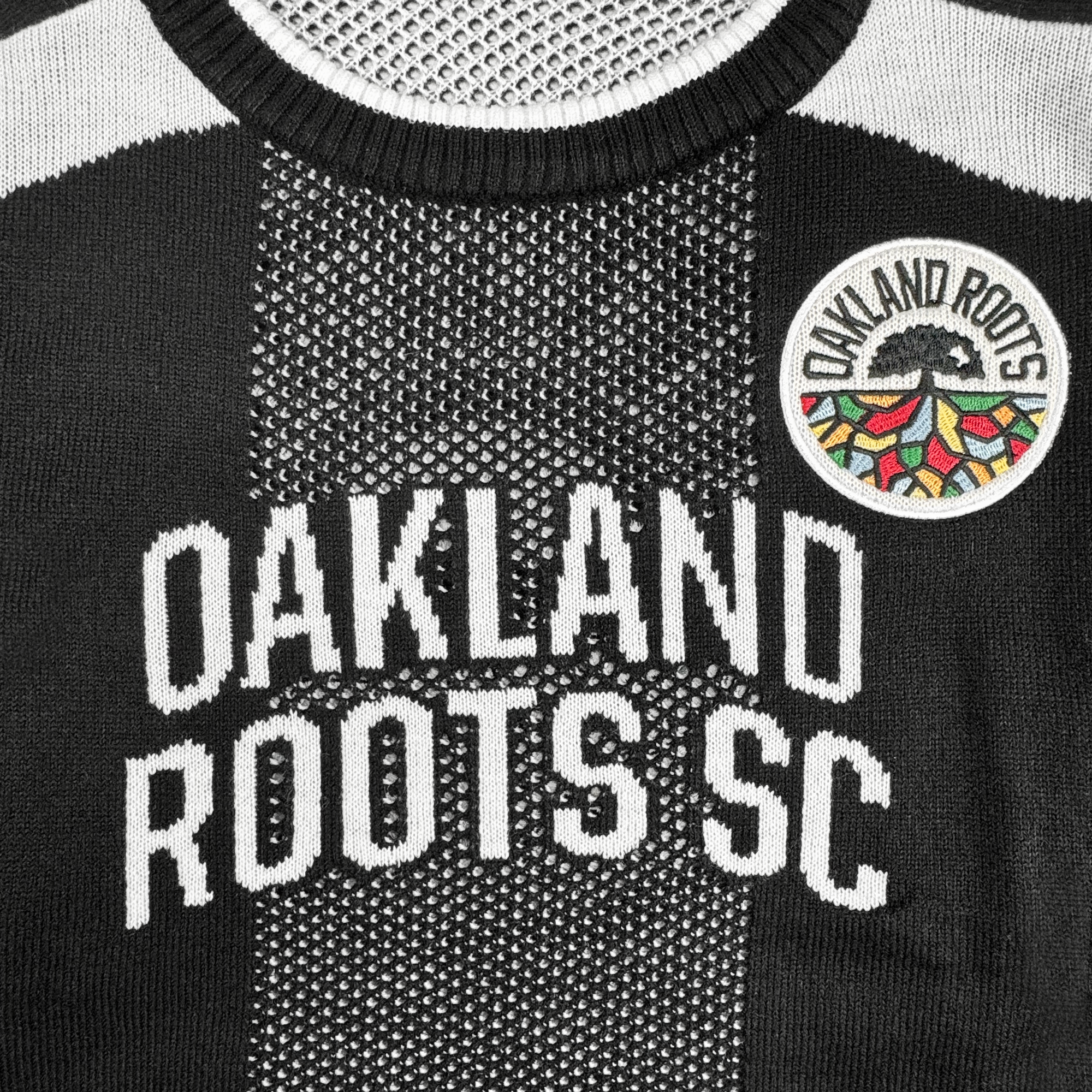 A close-up of the Oakland Roots SC Clubhouse Sweater by Oakland Roots SC, featuring "OAKLAND ROOTS SC" boldly across the chest and a vibrant patch on the right—classic style honoring this unique Oakland sports club.