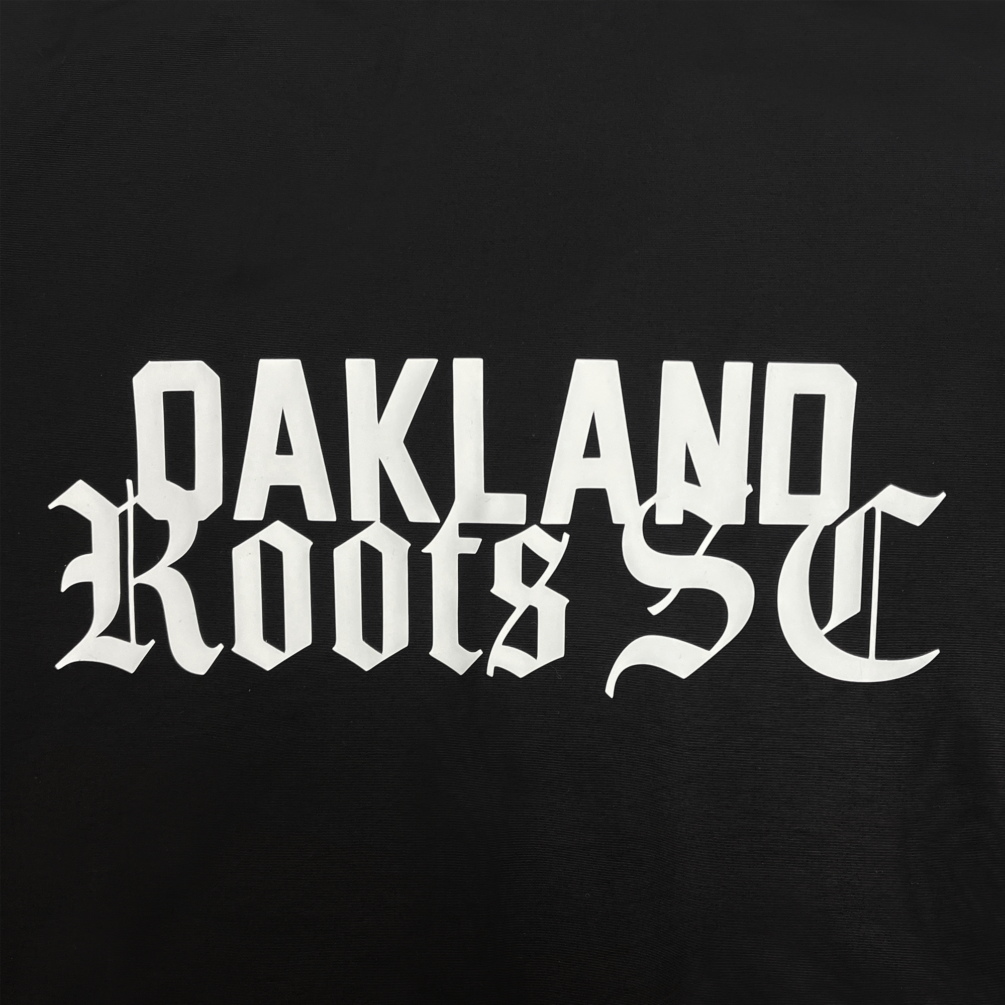 The Oakland Roots SC Coaches Jacket by Oakland Roots SC boasts a striking design with a black background, featuring bold, white Gothic font text that spells out "OAKLAND Roots SC." The central alignment places "OAKLAND" on the top line and "Roots SC" beneath it. This premium jacket merges style and water-resistant functionality seamlessly, making it a perfect choice for coaches.