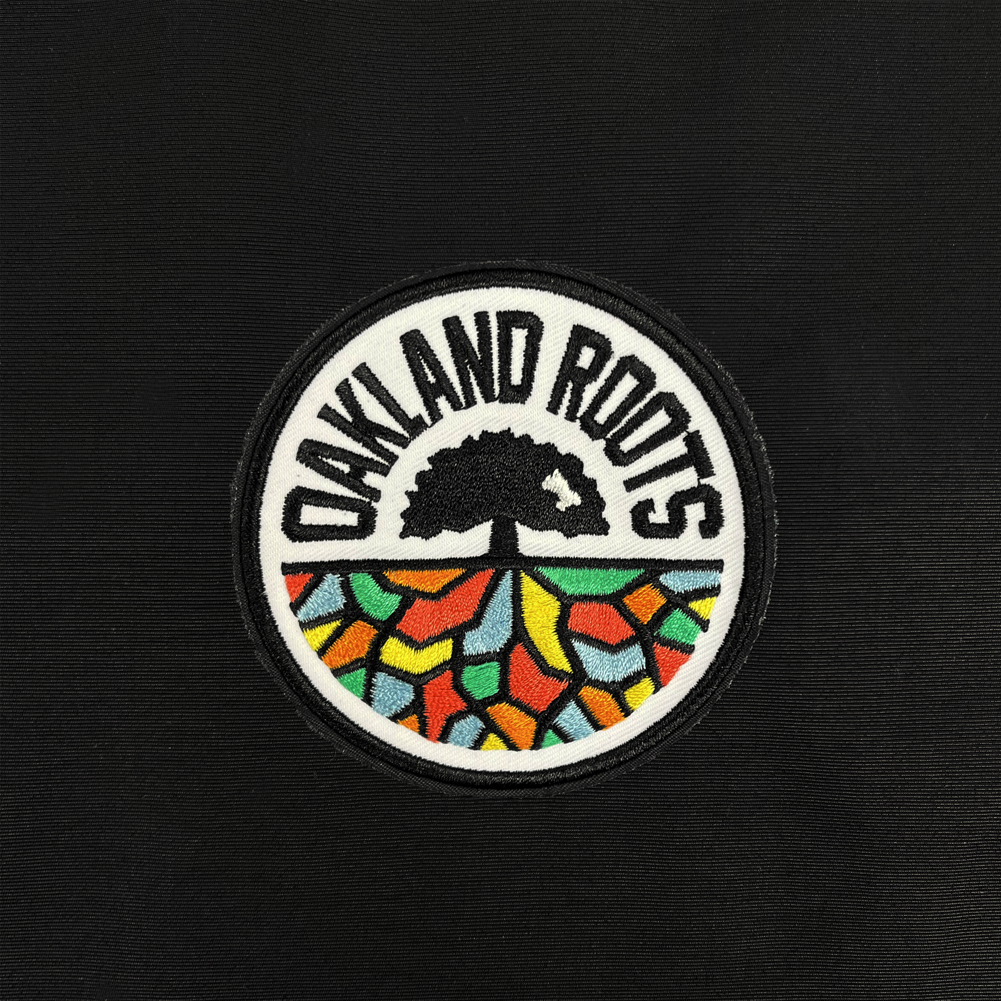 A black circular patch adorns the Oakland Roots SC Coaches Jacket, featuring a stylized tree with multi-colored geometric patterns in the lower half. The upper half showcases "OAKLAND ROOTS" in bold, white text curved around the top of the circle. This premium water-resistant jacket is ideal for any fan of Oakland Roots Sports Club.