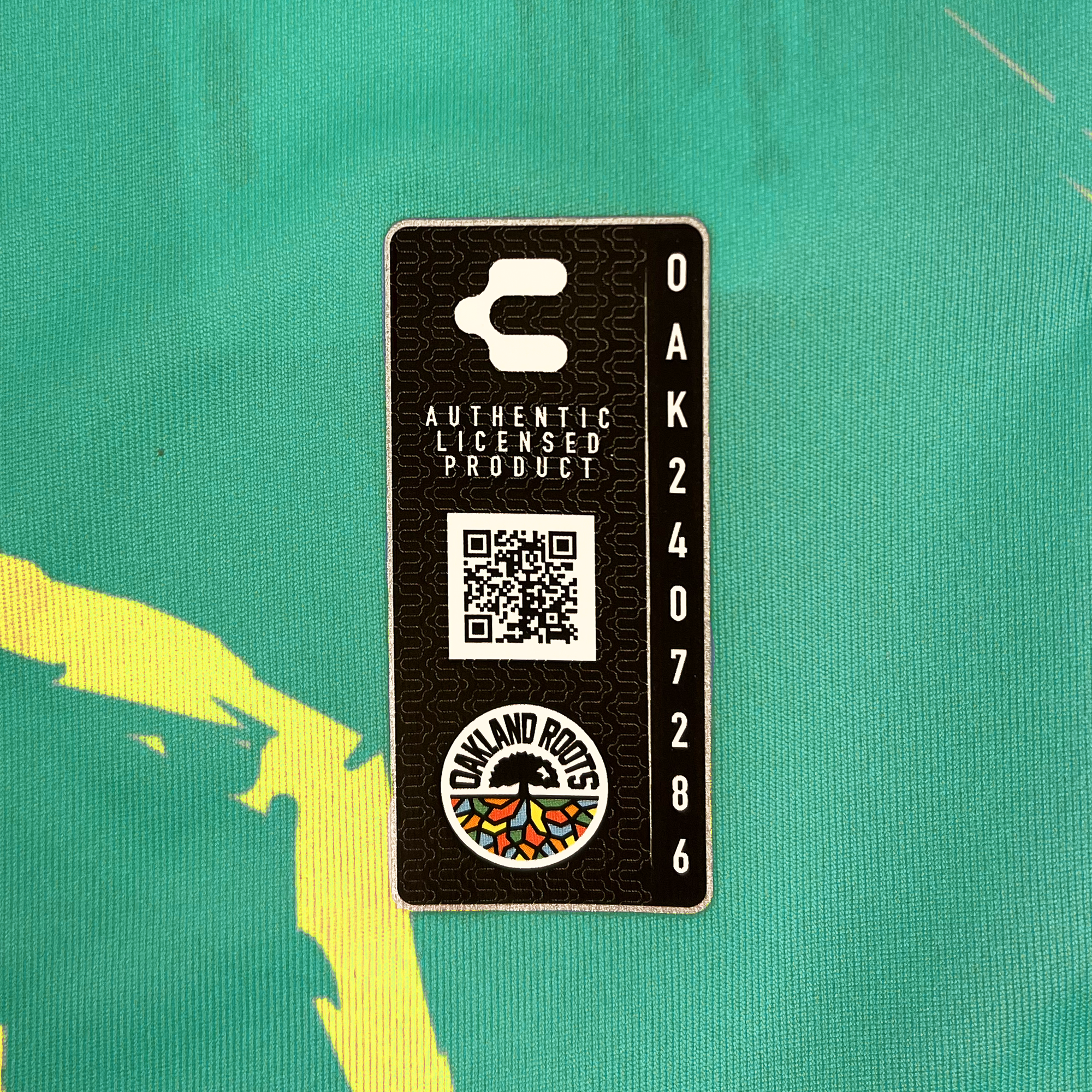 A rectangular tag with a black background, showcasing a QR code in the center and text that reads "Authentic Licensed Product" above it. Below the QR code is a circular logo featuring the text "Oakland Roots SC" and a vibrant graphic inspired by Oakland's colors. The code "OAK240786" runs vertically on the right side.
