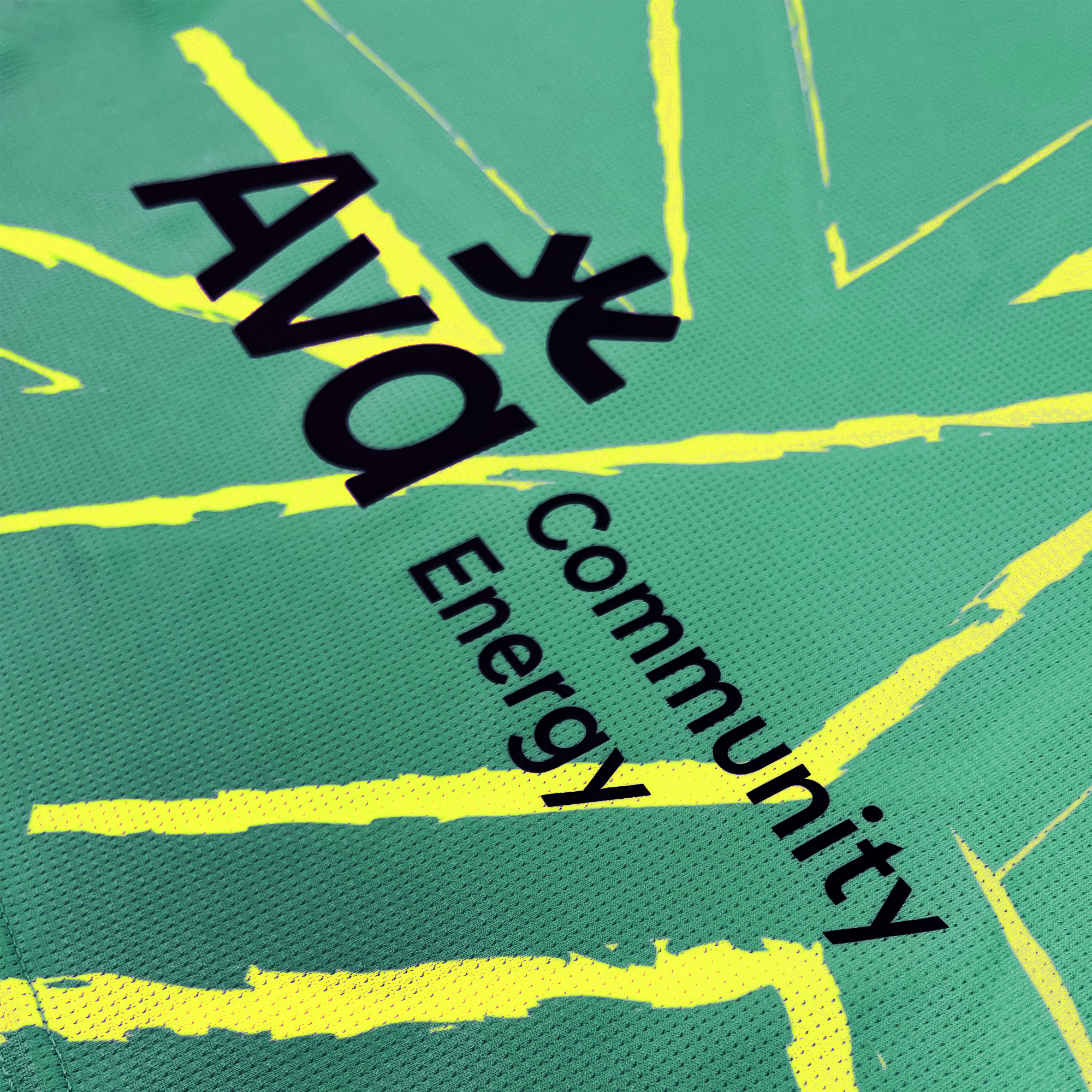 A green cloth with yellow lightning bolt designs and mosaic patterns, branded "Oakland Roots SC Goalkeeper Kit ft. Anthem Blue Cross." The words "AVA Community Energy" are printed in black on the fabric, along with a stylized abstract figure logo. The texture of the cloth appears to be mesh or breathable material, reminiscent of a traditional Goalkeeper Kit.