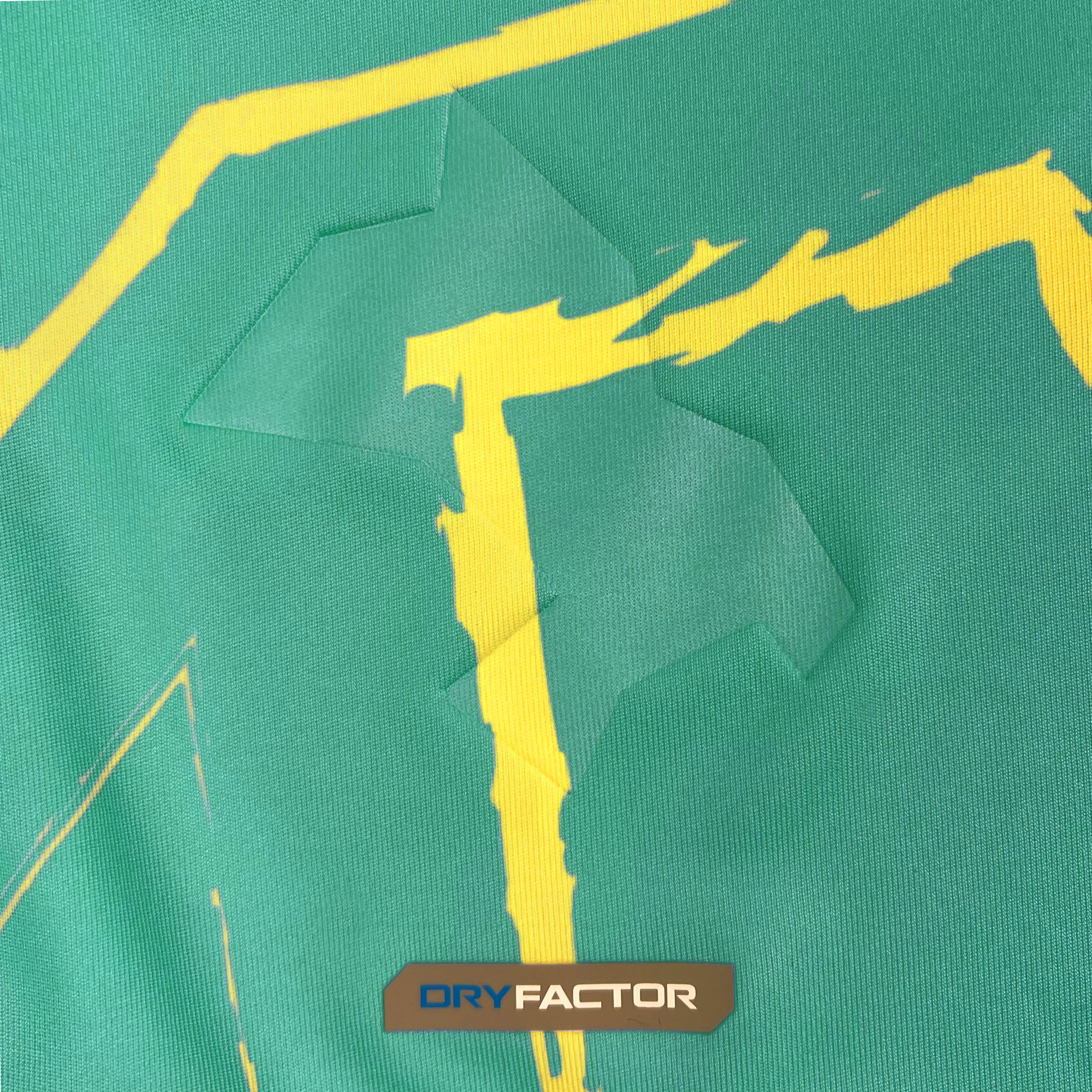 A close-up image of the fabric from the Custom Unisex Oakland Roots SC Goalkeeper Kit ft. Anthem Blue Cross, featuring an angular, jagged yellow pattern on a green background with visible creases. The lower part of the image showcases a gray rectangular label that reads "DRY FACTOR" in blue and white fonts. The smooth-textured fabric, designed by Oakland Roots SC, is perfect for a goalkeeper kit and displays slight wrinkles.