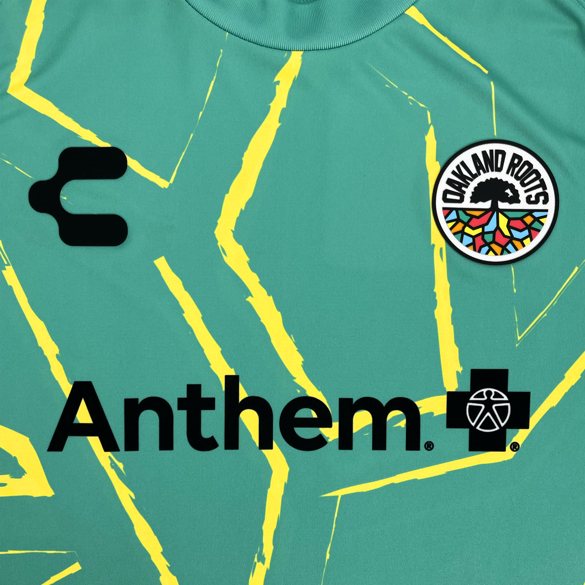 Introducing the Custom Unisex Oakland Roots SC Goalkeeper Kit featuring Anthem Blue Cross: A green sports jersey with jagged yellow lines, adorned with a black 'C' on the top left and the Oakland Roots emblem—a tree inside a circular badge with a colorful pattern and text—on the top right. The lower section showcases "Anthem" accompanied by a cross and an additional icon. Enhanced with DRY FACTOR technology, this jersey combines style and function.