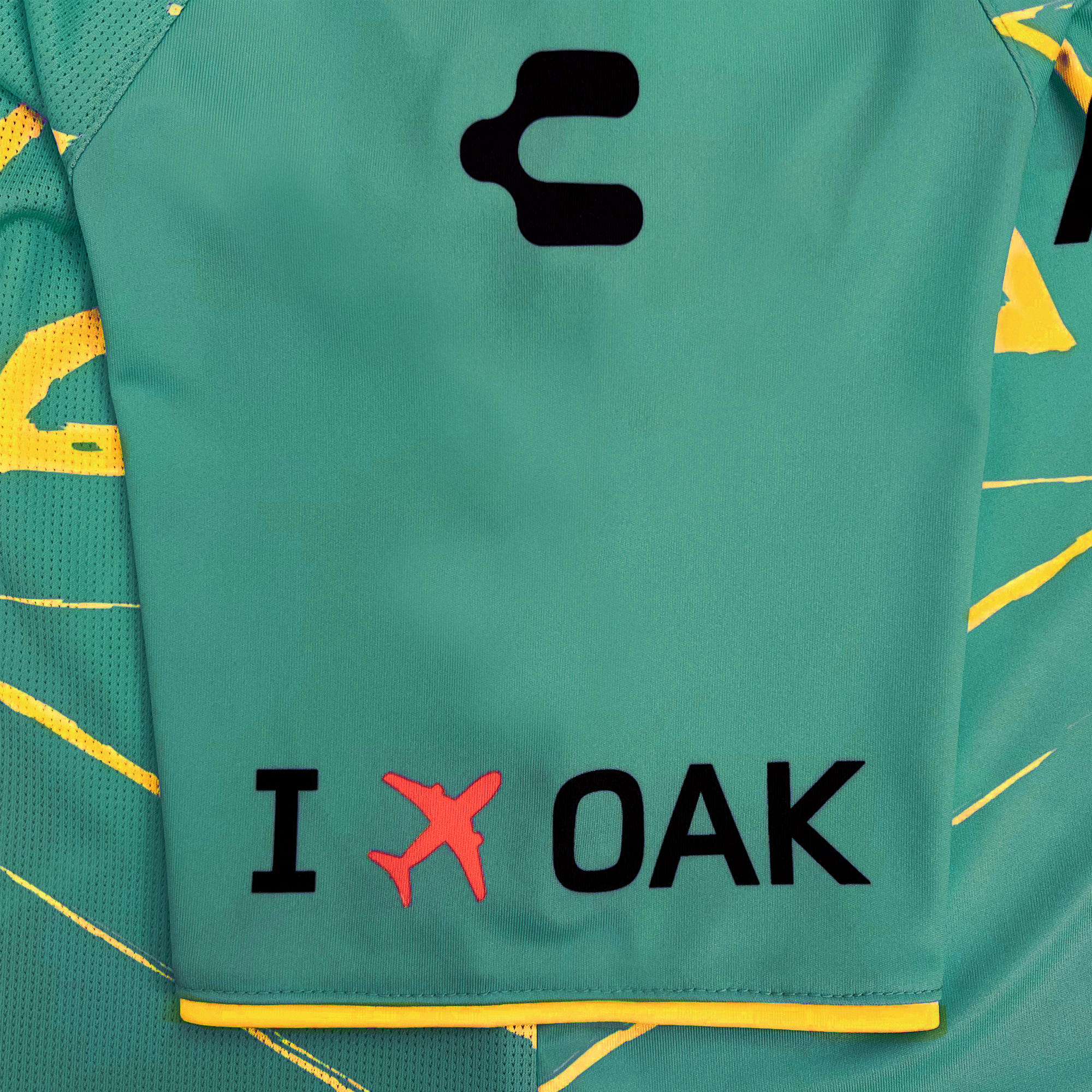 A close-up image of the Custom Unisex Oakland Roots SC Goalkeeper Kit ft. Anthem Blue Cross reveals a logo resembling the letter 'C' in black. Below, black text reads "I *airplane icon* OAK", with the airplane icon in red. Yellow accents and abstract graphic patterns in yellow are visible on the jersey's background, showcasing DRY FACTOR technology from Oakland Roots SC.