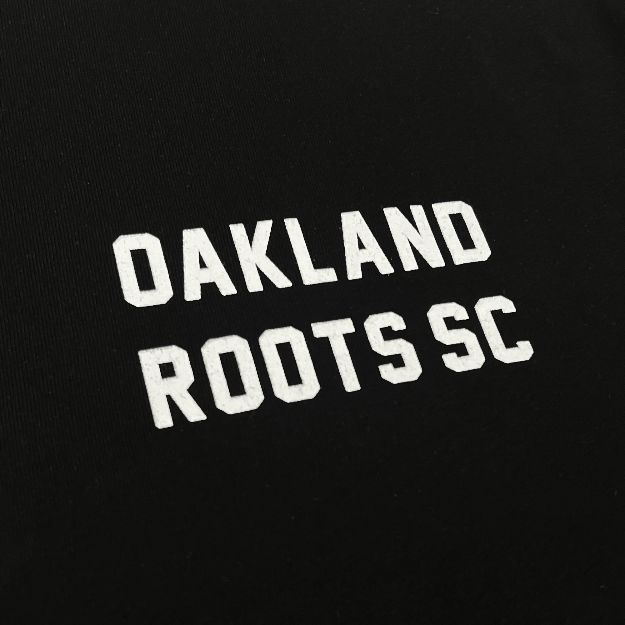 Close-up of the OAKLAND ROOTS SC wordmark on the chest of a black long-sleeve t-shirt.