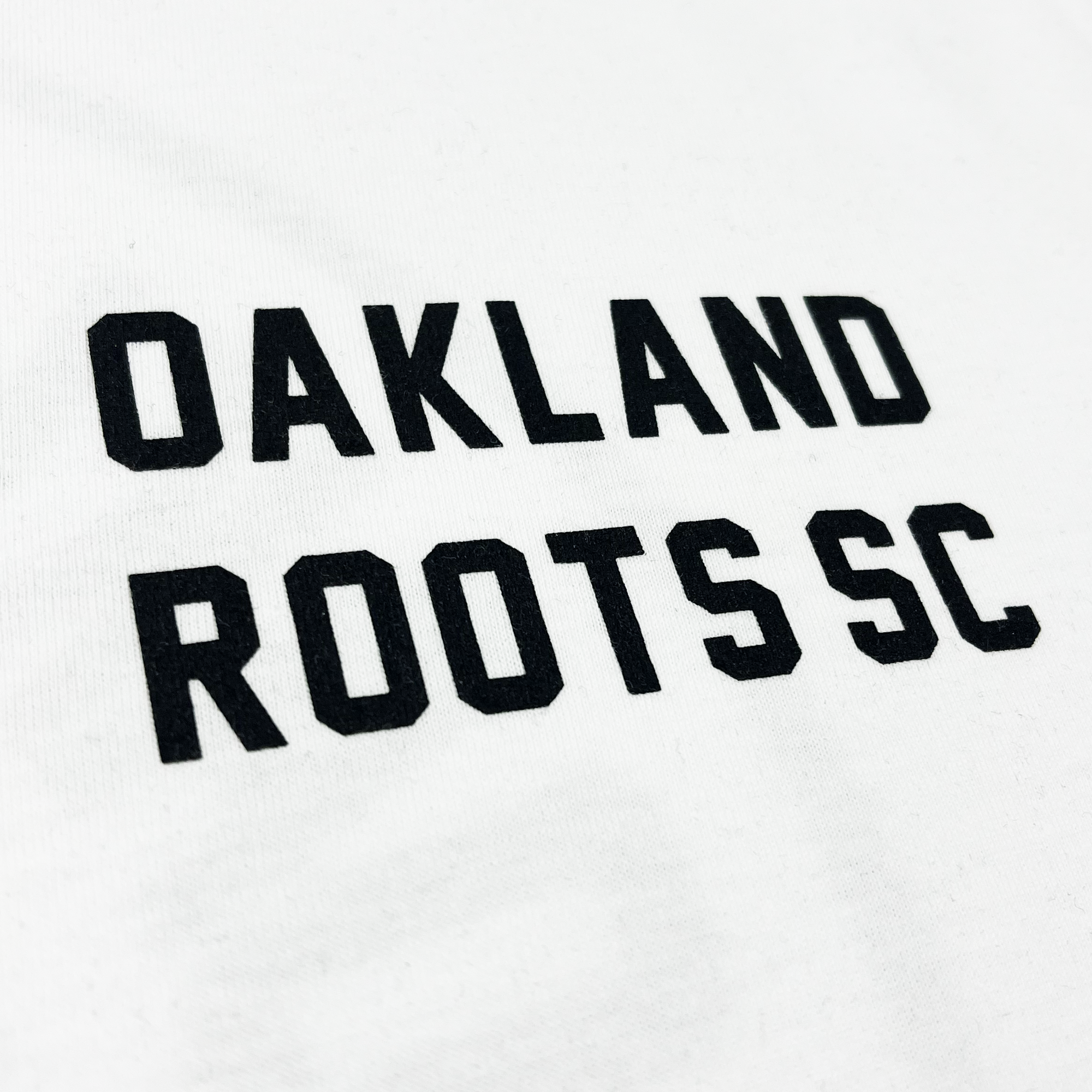 Close-up of the OAKLAND ROOTS SC wordmark on the chest of a white long-sleeve t-shirt.