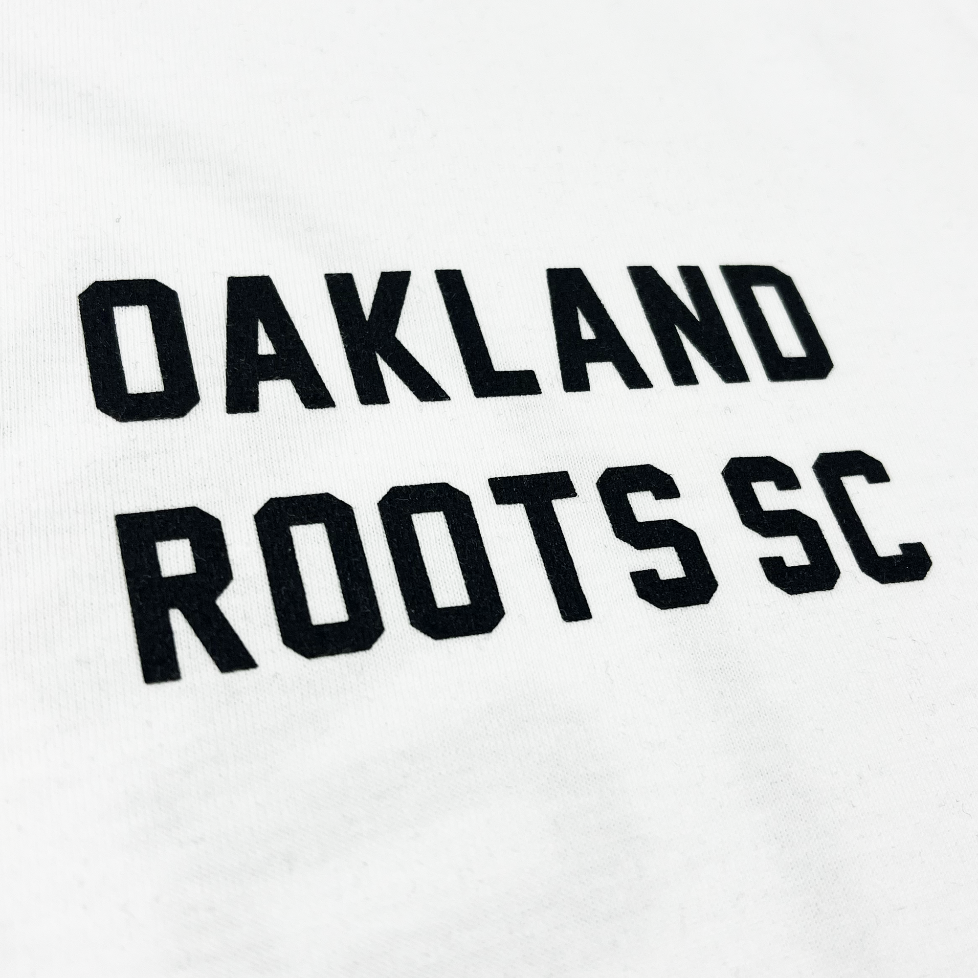 The Oakland Roots SC Classic 2.0 LS Tee features a white long sleeve design with bold, black text centered on it. The clean and modern font displays "OAKLAND ROOTS" in all capital letters on the top line and "SC" on the bottom line, celebrating the sports club's charm.