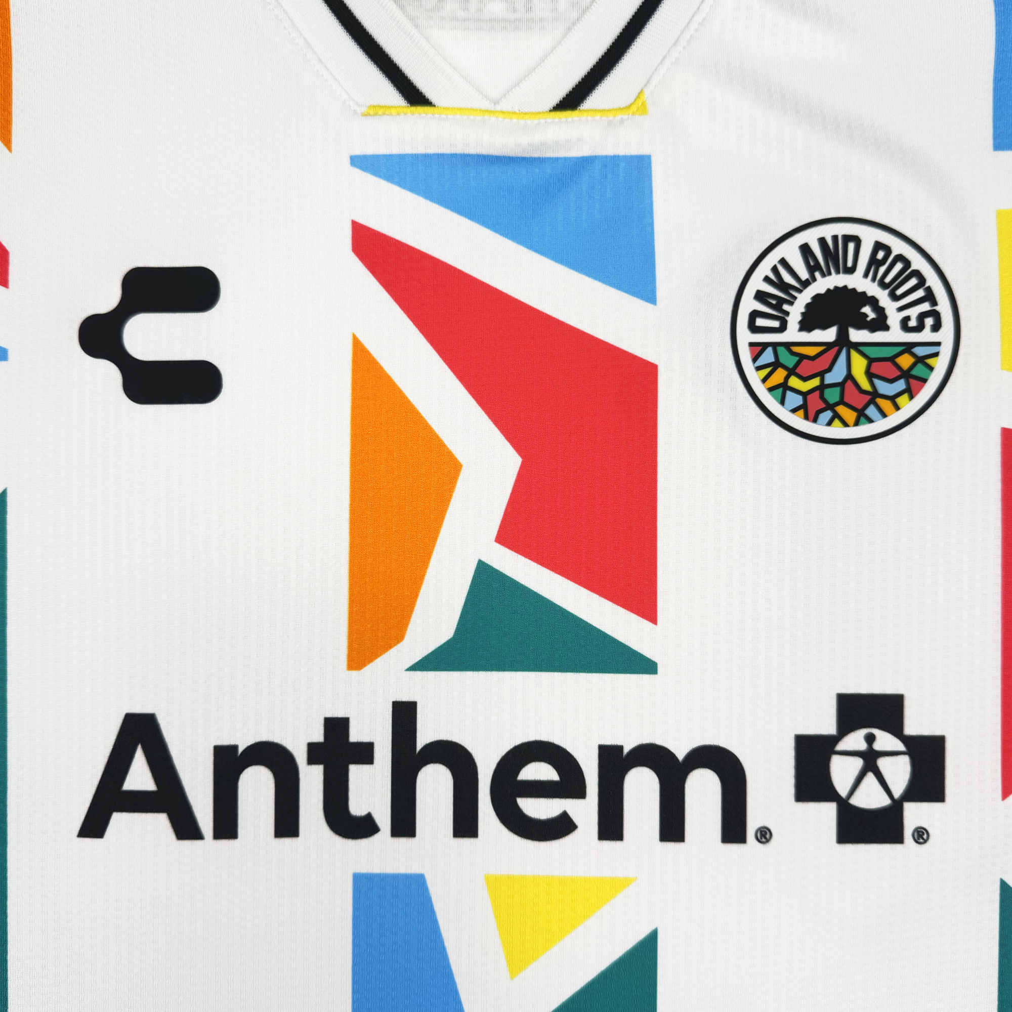 Close-up of the Custom Unisex Mosaic in Motion Kit by Oakland Roots SC: a white jersey featuring colorful geometric shapes, custom details, the iconic tree logo, bold “Anthem” text with cross, C-shaped emblem, and a striped black-and-white collar.