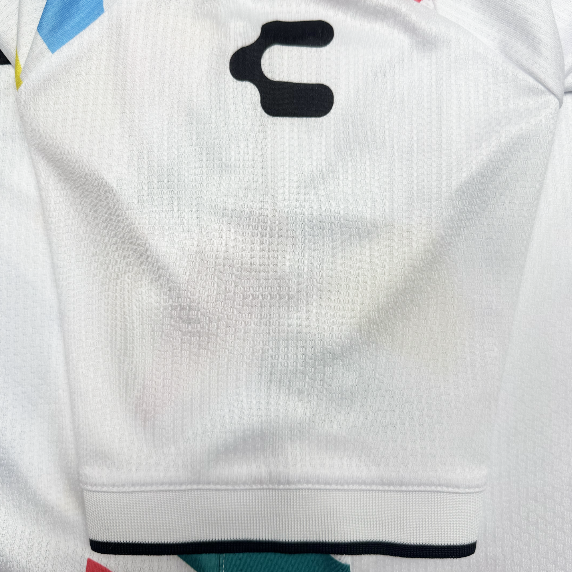 A close-up of the Custom Unisex Mosaic in Motion Kit by Oakland Roots SC shows a white sleeve with a bold black "C" near the shoulder, bordered by black piping at the hem, with hints of colorful mosaic patterns in the background.