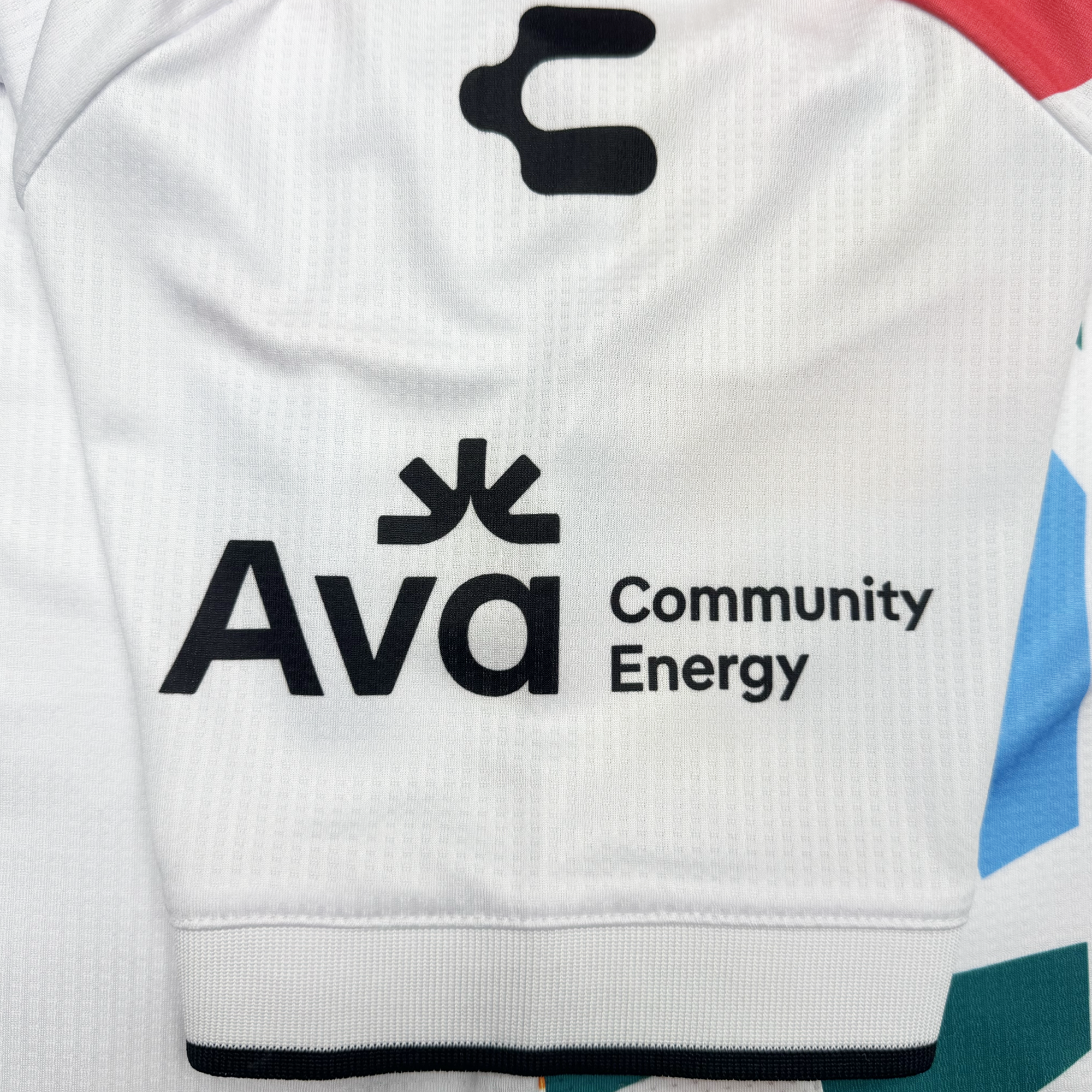 A close-up of the Unisex Mosaic in Motion Kit by Oakland Roots SC shows a white mosaic jersey sleeve with the black “Ava Community Energy” logo and stylized symbol, plus colorful geometric shapes from the 2026 kit on a partially visible sleeve.