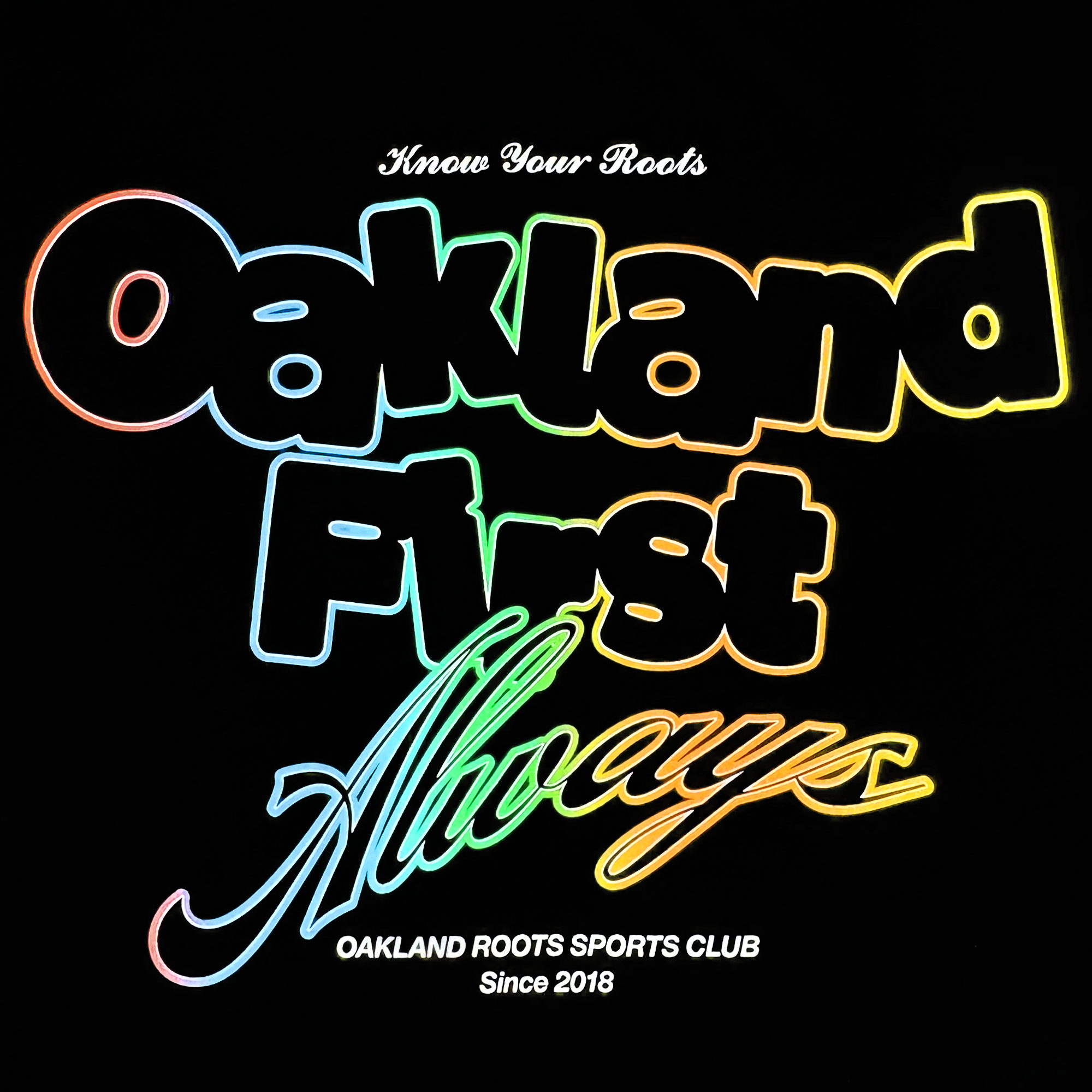 The image depicts the "OFA Different Tee" featuring "Oakland First Always" in bold, multicolored neon letters. Above, "Know Your Roots" appears in small white script and below is "Oakland Roots Sports Club Since 2018," representing Oakland Roots SC's dedication to sports and social good on a black background.
