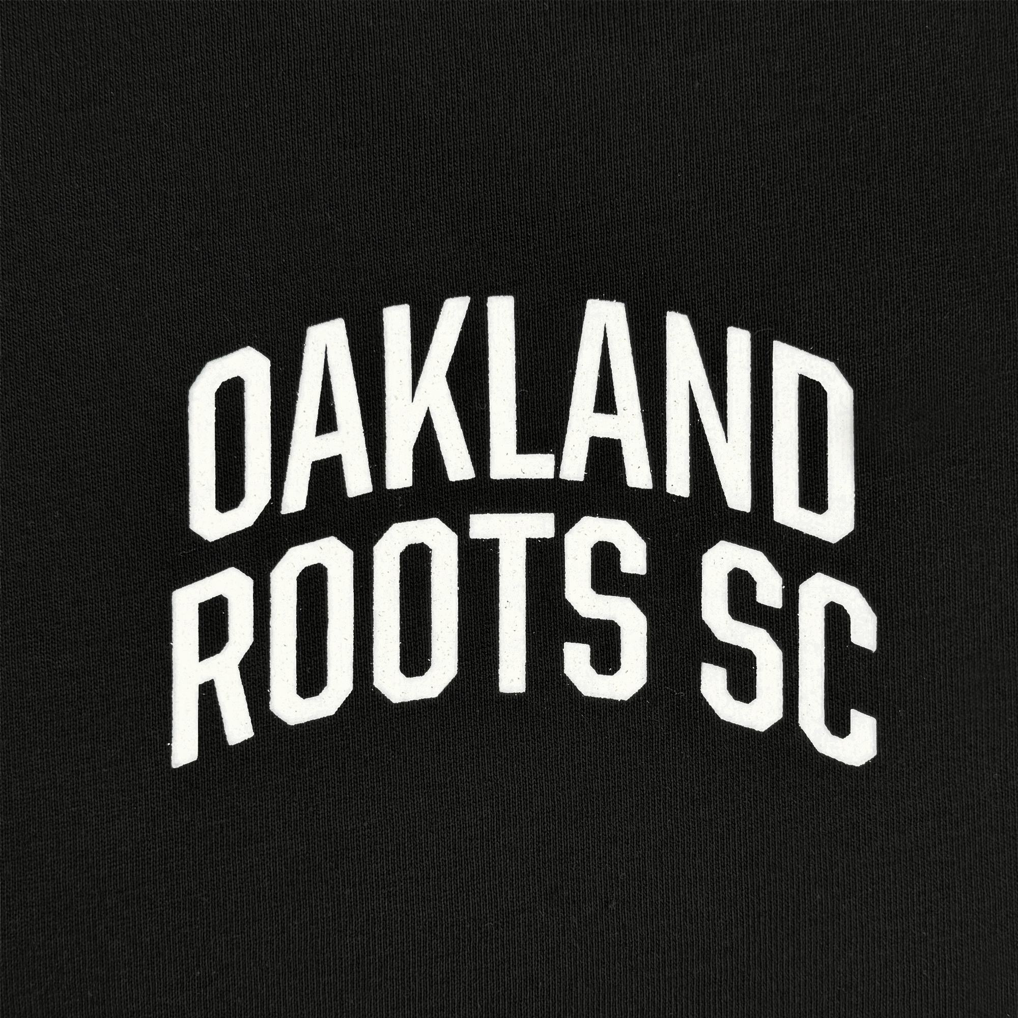 The OFA Worldwide Tee from Oakland Roots SC features bold, white capital letters "OAKLAND ROOTS SC" arching across a black backdrop, representing the club's dedication to social good.