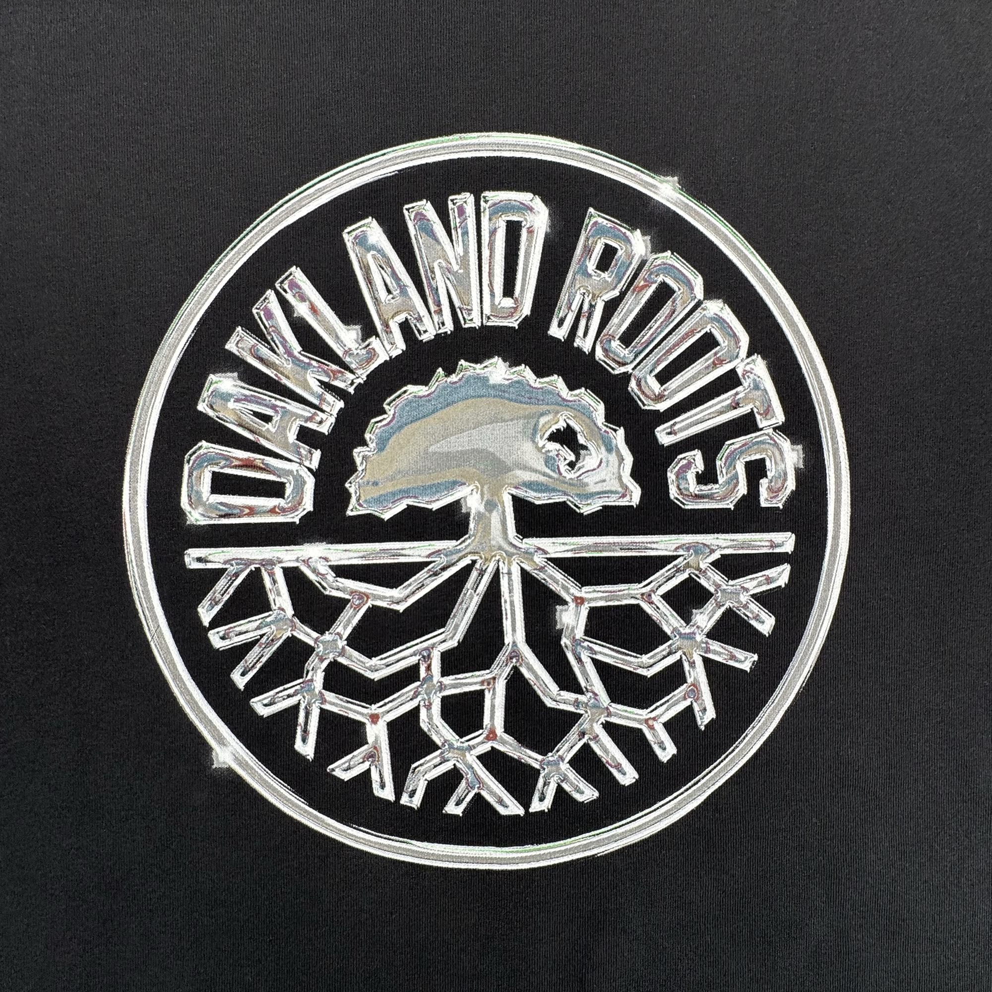 The Platinum Crest Tee by Oakland Roots SC features a circular silver logo with an oak tree and roots on a black background, with "OAKLAND ROOTS" curved above—a standout design for fans and the perfect classic fit t-shirt.