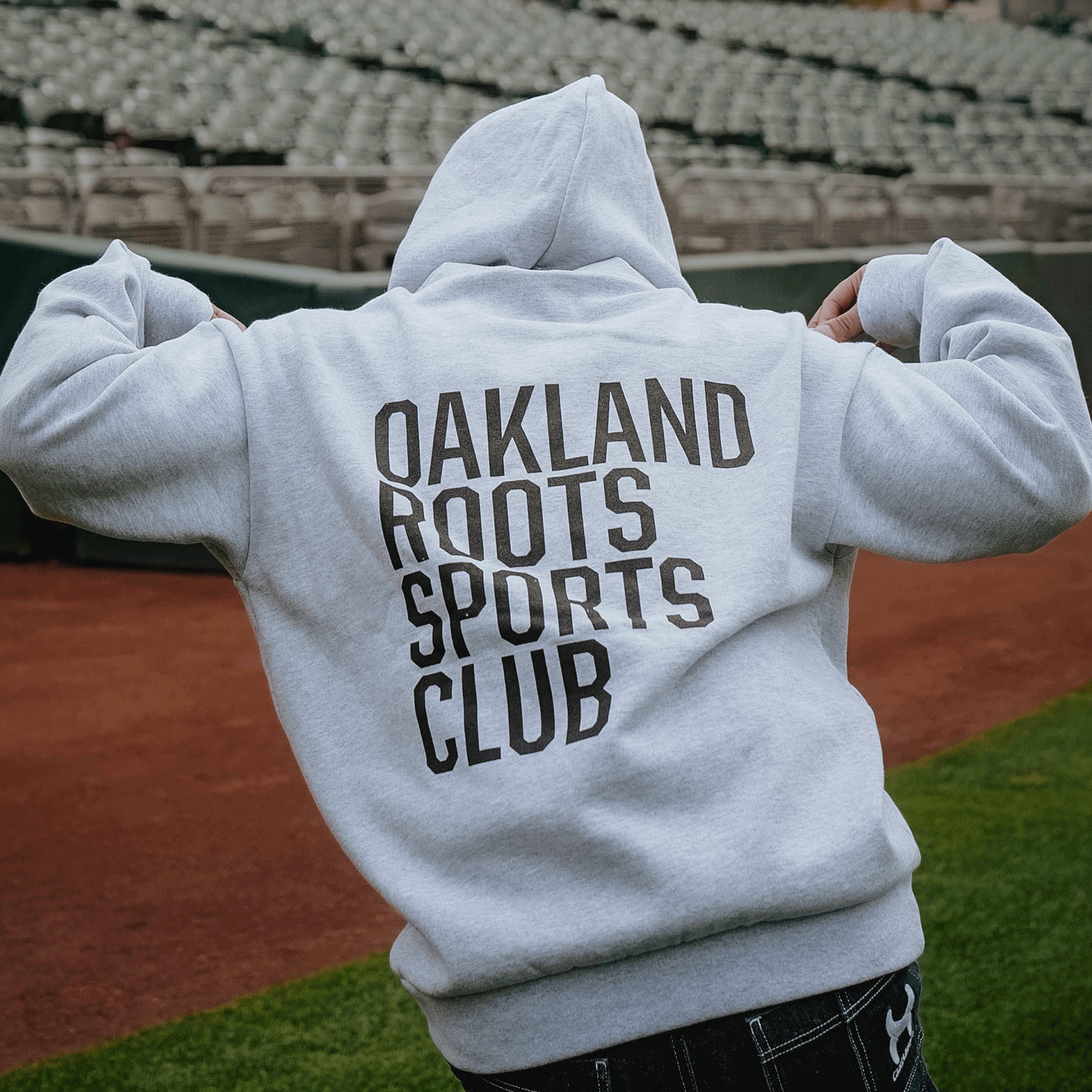 Wearing the Oakland Roots SC Heavy Hoodie by Oakland Roots SC, a person stands on a sports field facing empty stadium seats, hood up and hands at their sides, with bold “OAKLAND ROOTS SPORTS CLUB” text visible on the back.