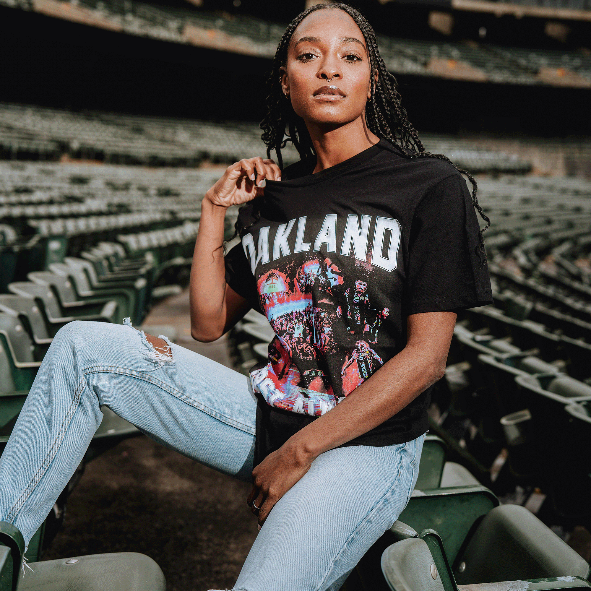 A woman with braided hair sits on green stadium seats, confidently showing off her black OFA Tour Tee by Oakland Roots SC, paired with ripped jeans as she faces the camera and proudly displays her love for the club.