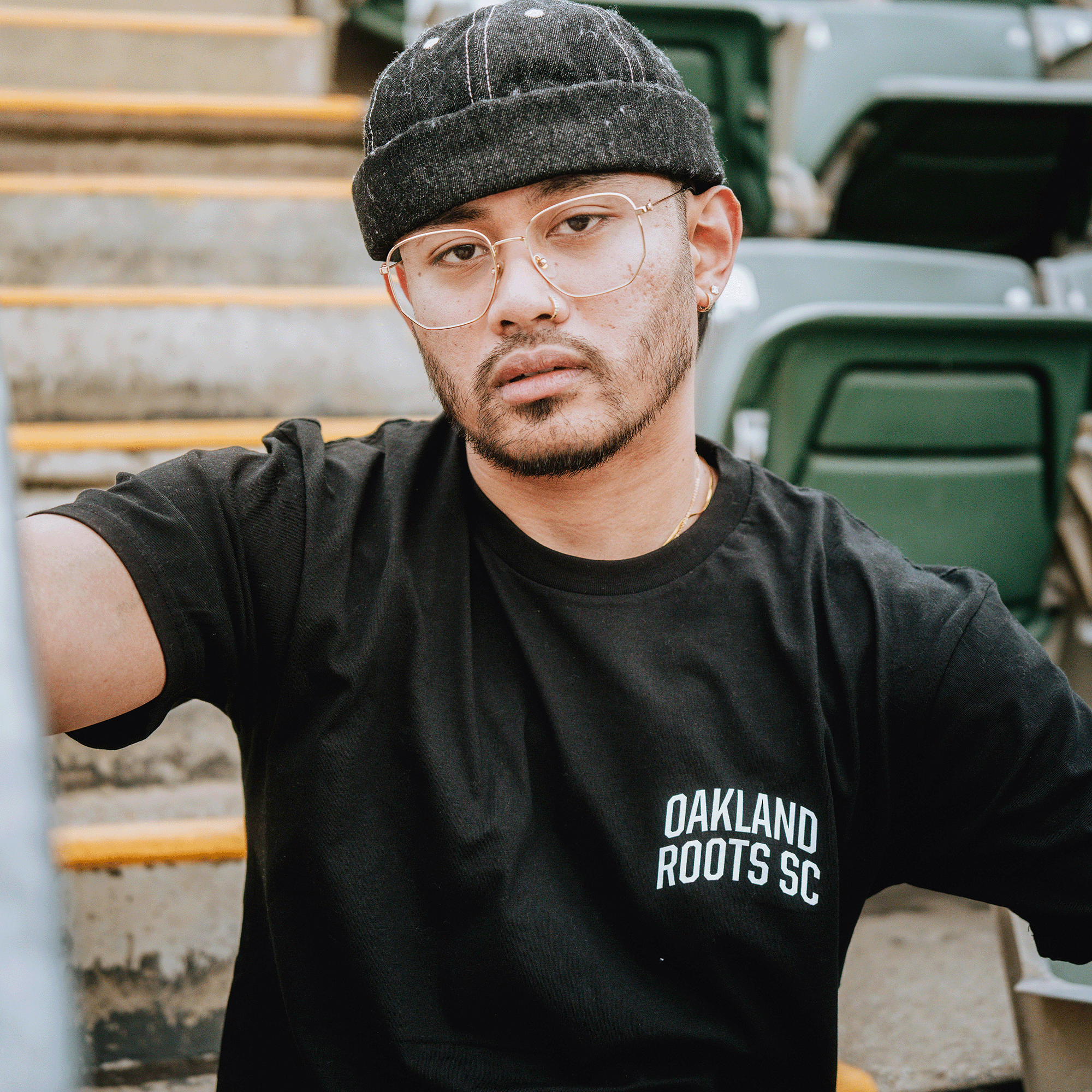 A man with glasses, a black beanie, and a trimmed beard sits on stadium steps with green seats behind him, wearing the OFA Worldwide Tee by Oakland Roots SC. He looks directly at the camera with a neutral expression.