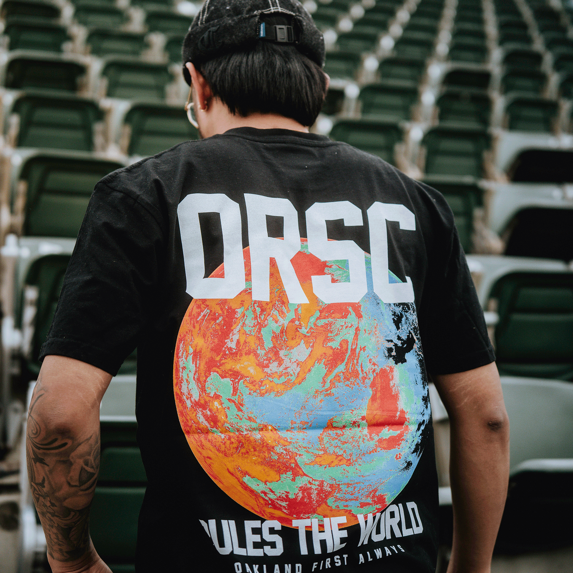 A person with a tattooed arm and black cap faces away in empty stadium seats, wearing the OFA Worldwide Tee by Oakland Roots SC—a black T-shirt featuring a colorful Earth graphic and bold "ORSC" lettering.