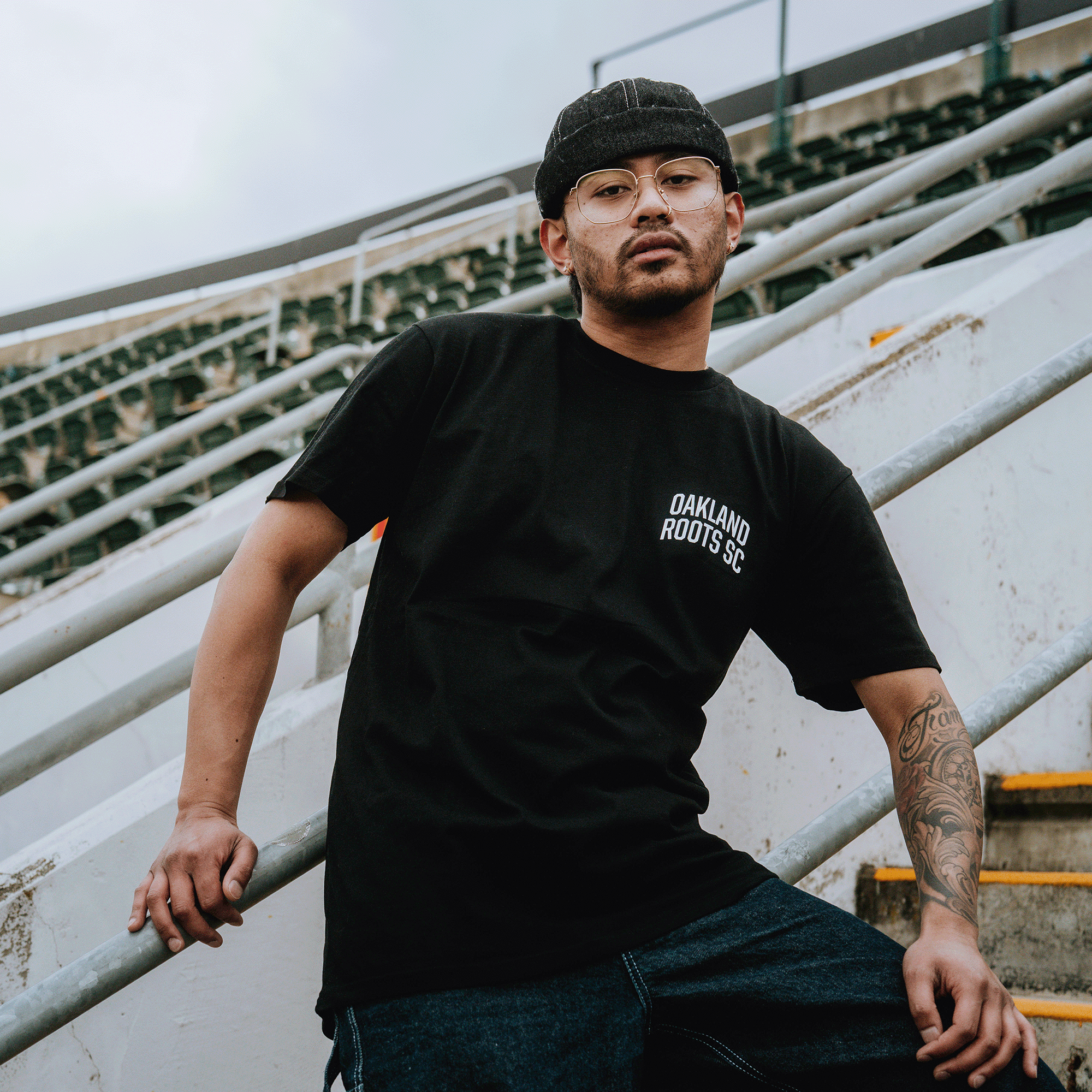 A man with glasses and a backward cap leans on a stadium stair railing, wearing the Oakland Roots SC OFA Worldwide Tee. A tattoo is visible on his forearm, with empty concrete steps and metal railings in the background.