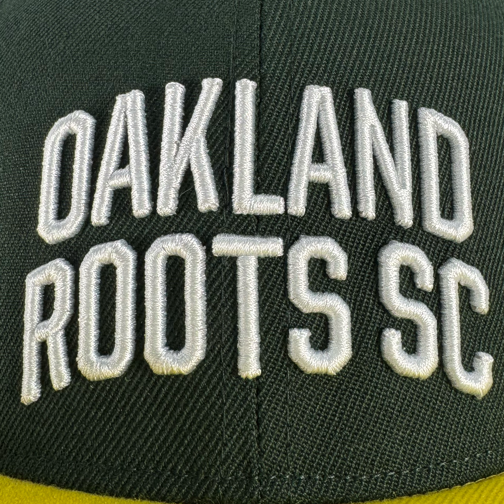 A close-up of the Oakland Roots SC Arch Text Snapback by Oakland Roots SC, featuring large white embroidered "OAKLAND ROOTS SC" text, visible fabric texture and stitching, and a hint of yellow at the bottom edge.
