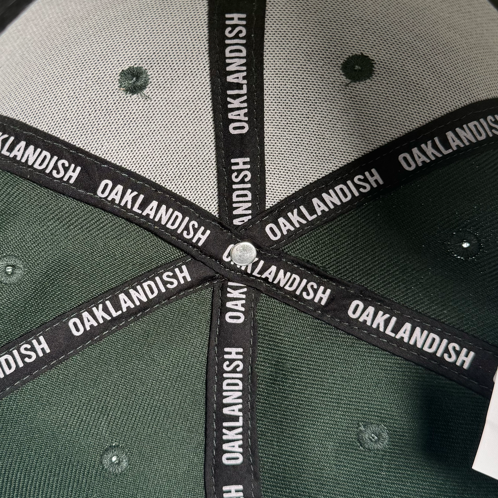 Close-up of the Oakland Roots SC Arch Text Snapback's dark green interior top. Black fabric strips reading "OAKLANDISH" in white divide the panels. White stitching is visible—perfect for any Oakland Roots SC fan.