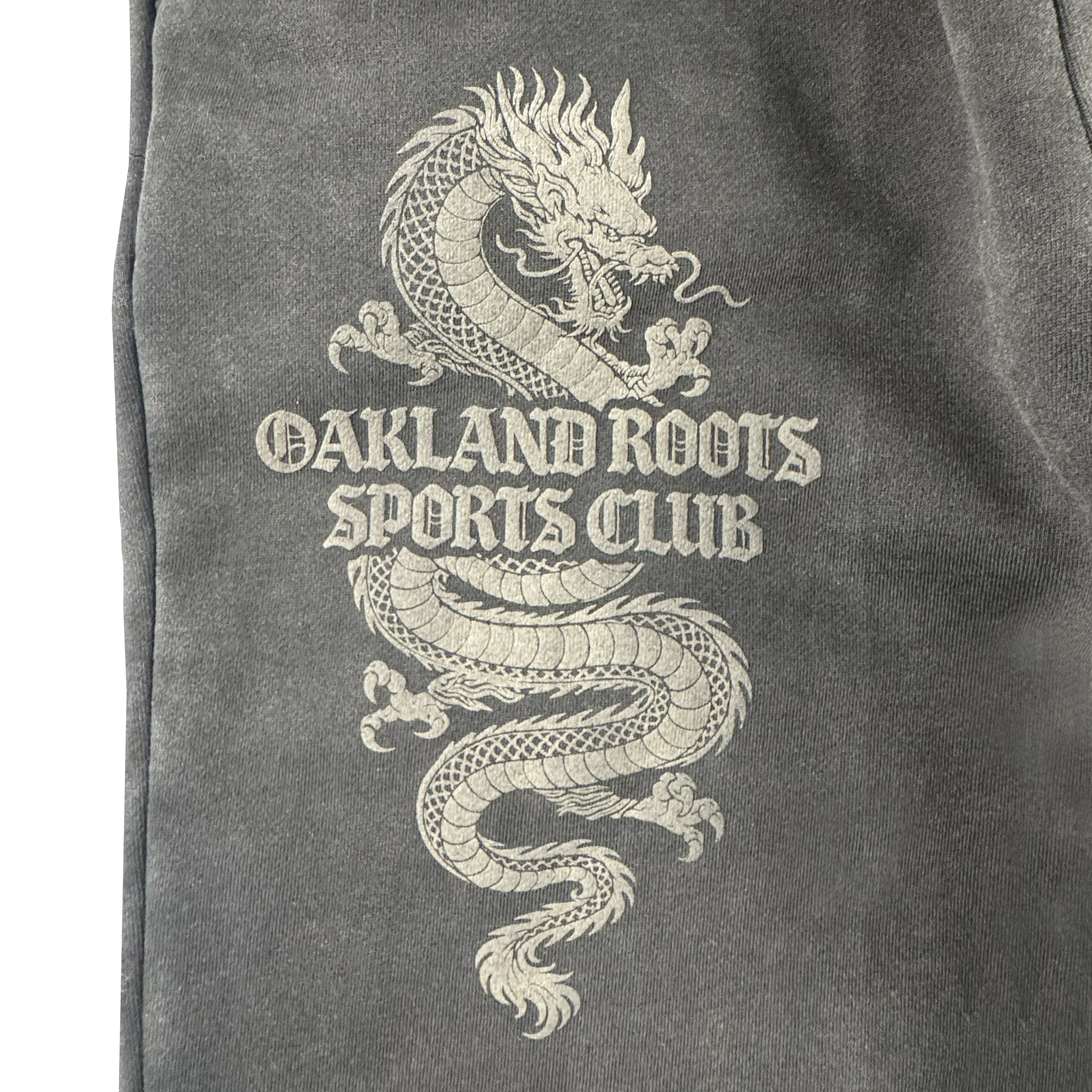 A detailed white dragon coils around bold, Gothic “OAKLAND ROOTS SC” text on dark gray organic cotton Tatsu Pants, capturing Oakland Roots pride.