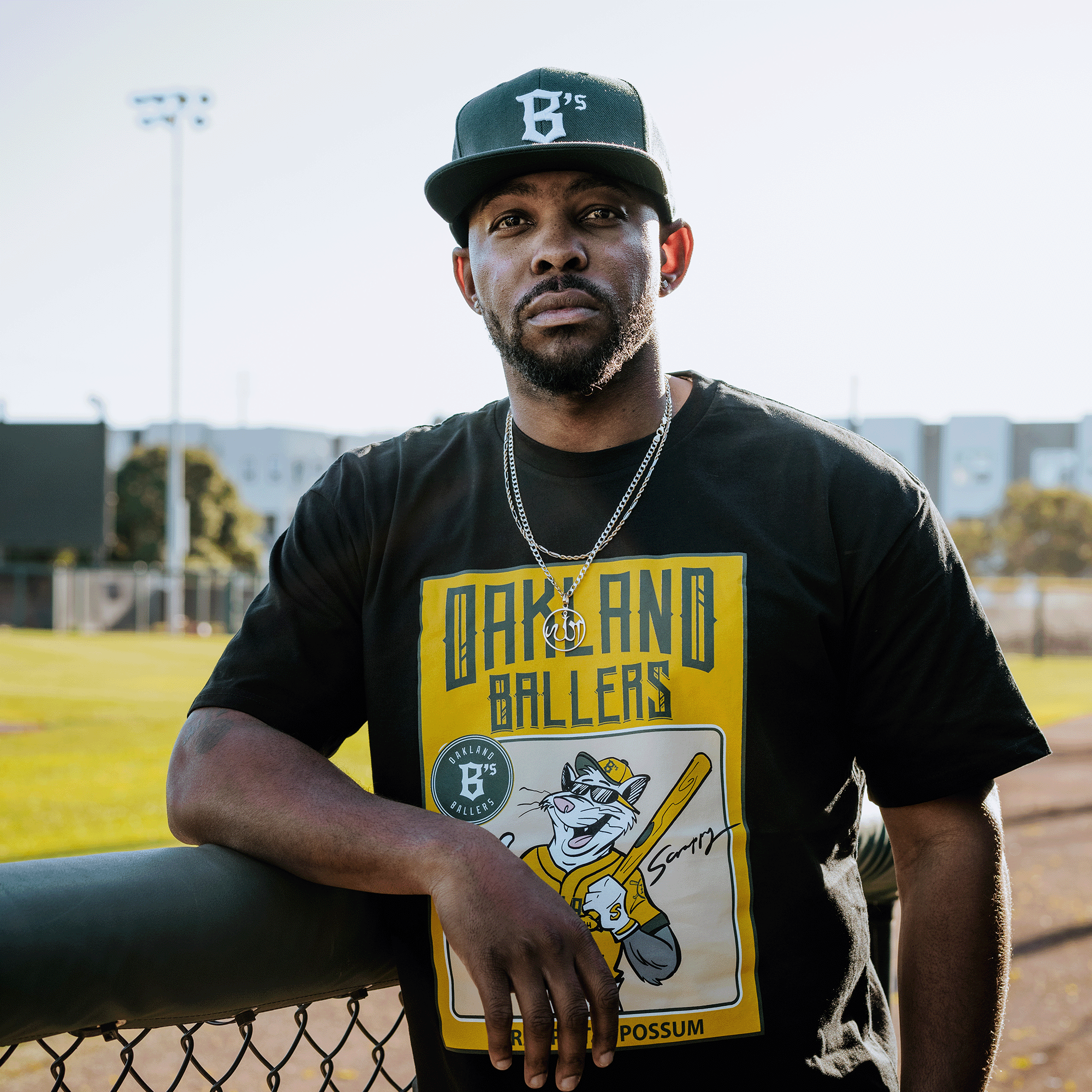 At Raimondi Park, a man leans on a chain-link fence by the baseball field, wearing a black Scrappy Rookie Card Tee and cap from Oakland Ballers featuring Scrappy the Rally Possum with a bat. In the background are partial bleachers under clear skies.