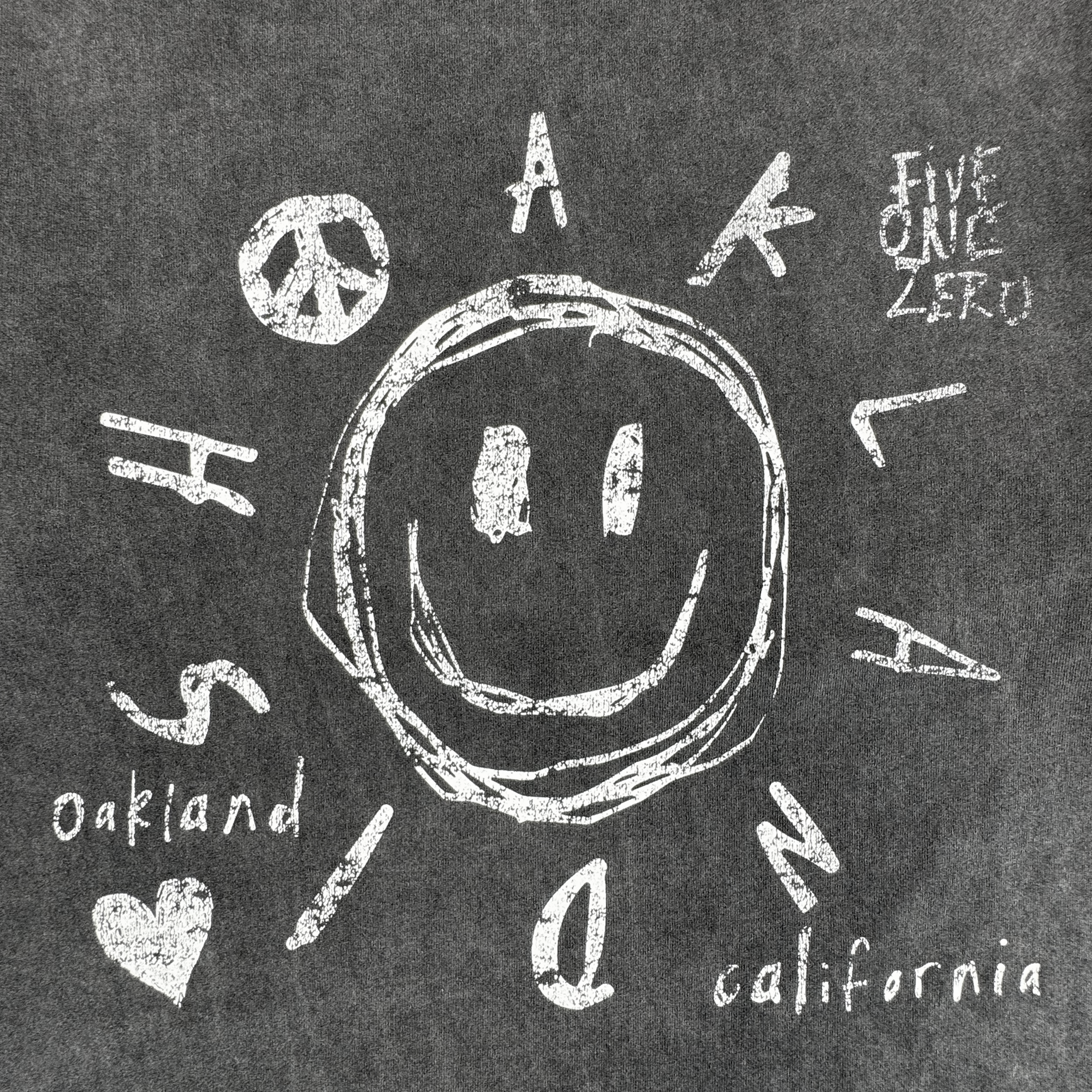 A chalk-like graphic on a vintage black Scribble LS Tee by Oaklandish features a big smiley face, playful "OAKLAND" text, "oakland," "california," "FIVE ONE ZERO," and a peace sign. 100% cotton classic fit.