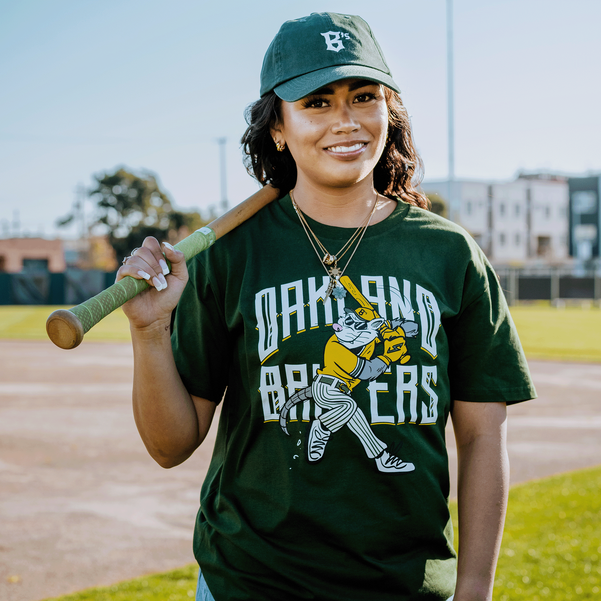 A woman wearing a green cap and an "Oakland Ballers" Slugging Scrappy Tee stands on a baseball field at Raimondi Park. Her hair is down, adorned with gold necklaces, as she holds a bat over her shoulder and smiles with buildings visible in the background.