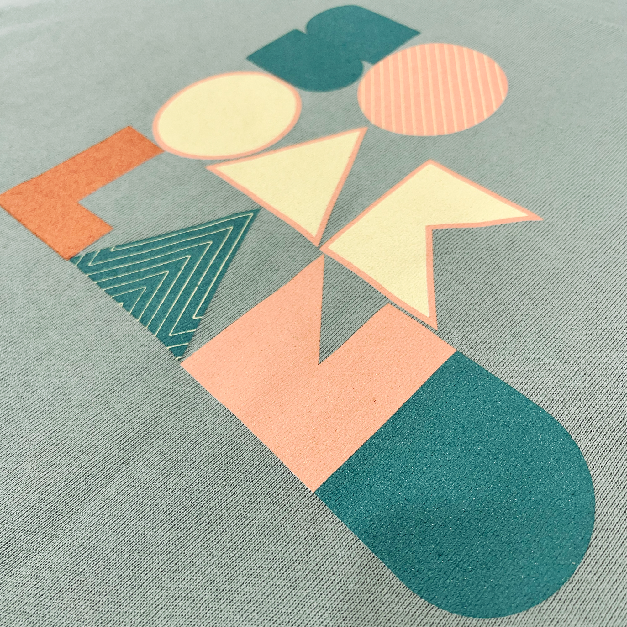 Detailed close up of full-color SOOAKLAND graphic wordmark on the front chest of a cropped crewneck sage green sweatshirt.