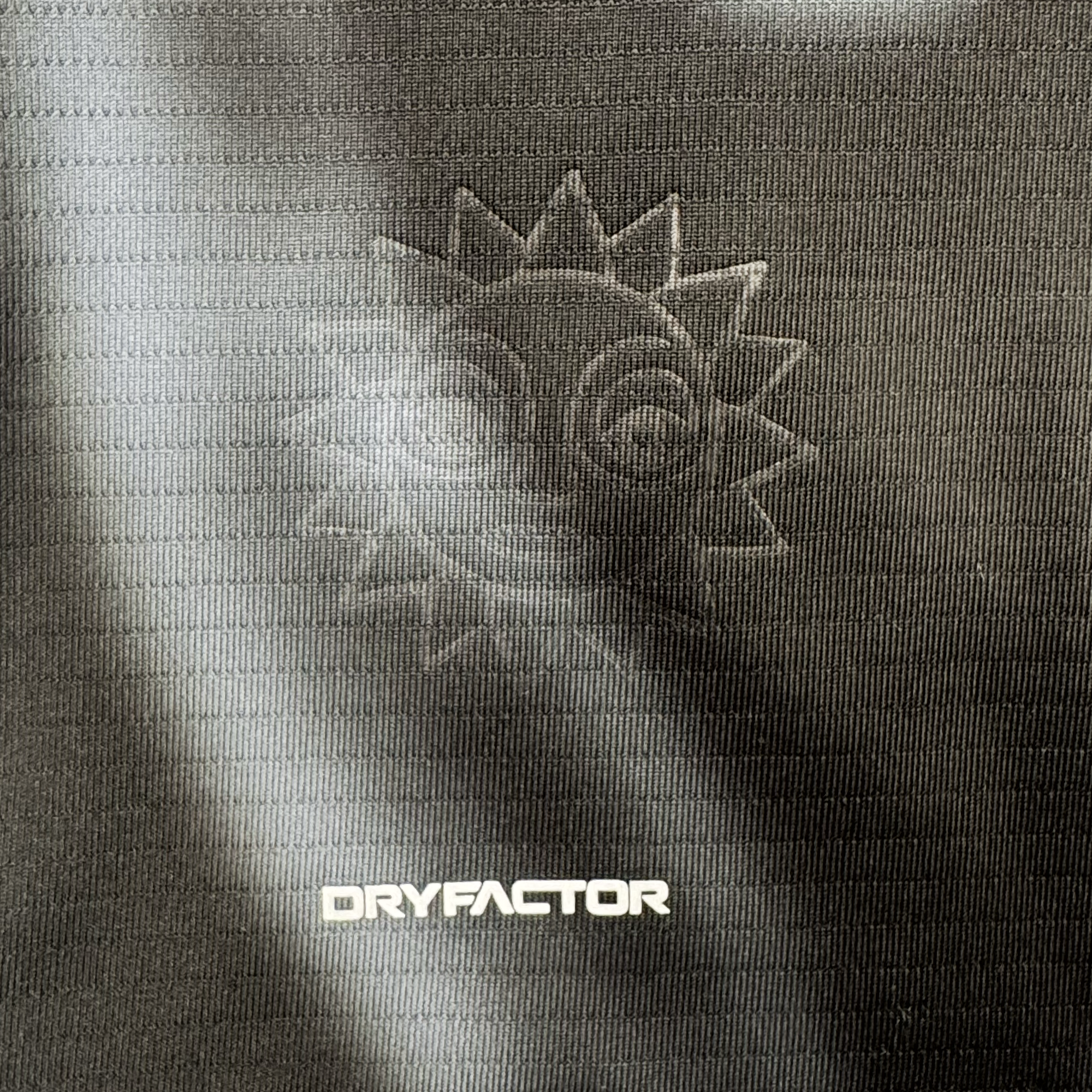A close-up of the Custom 2026 OAKLAND SOUL SC SPIRIT JERSEY fabric by Oakland Soul shows a subtle embossed sun with a smiling face above the white “DRYFACTOR” logo, textured dark material with horizontal lines, and soft curved light reflections.