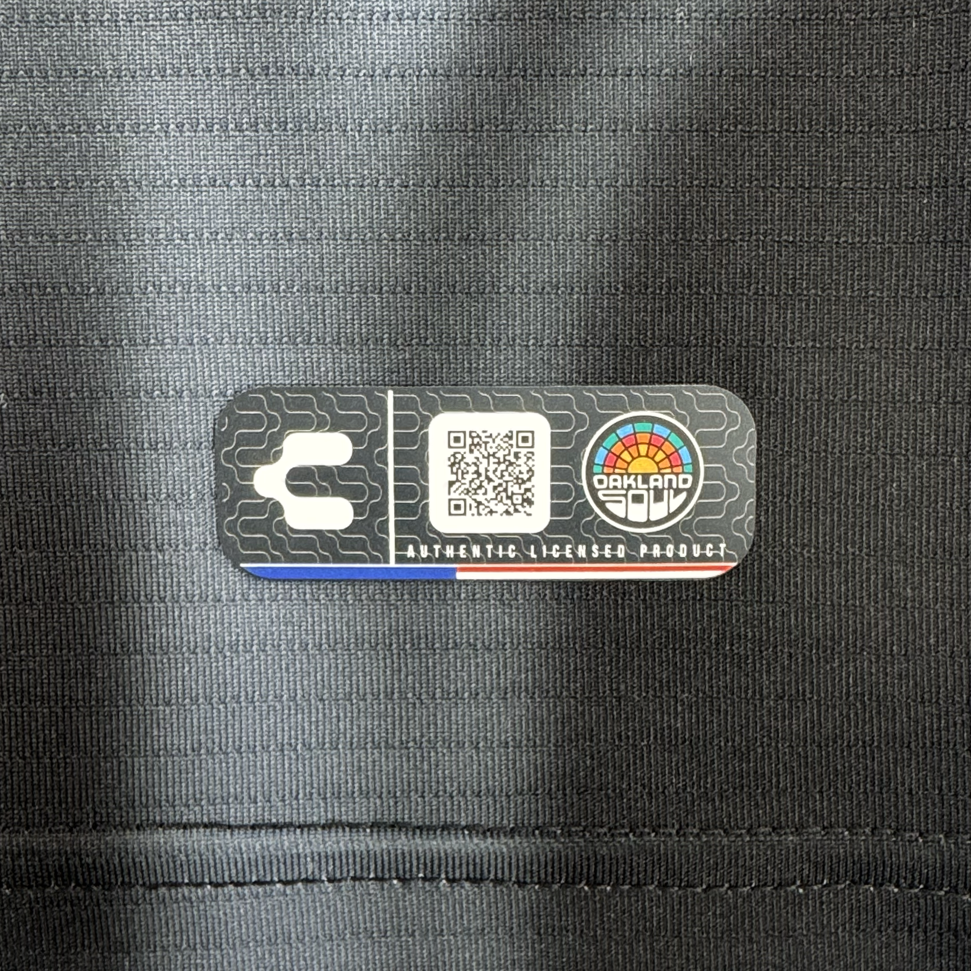 A close-up of the black fabric tag on the 2026 OAKLAND SOUL SC SPIRIT JERSEY by Oakland Soul shows a QR code, Charly’s "C" logo, a round rainbow "OAKLAND" logo, and a polyester tag reading "AUTHENTIC LICENSED PRODUCT" with a subtle grid pattern.