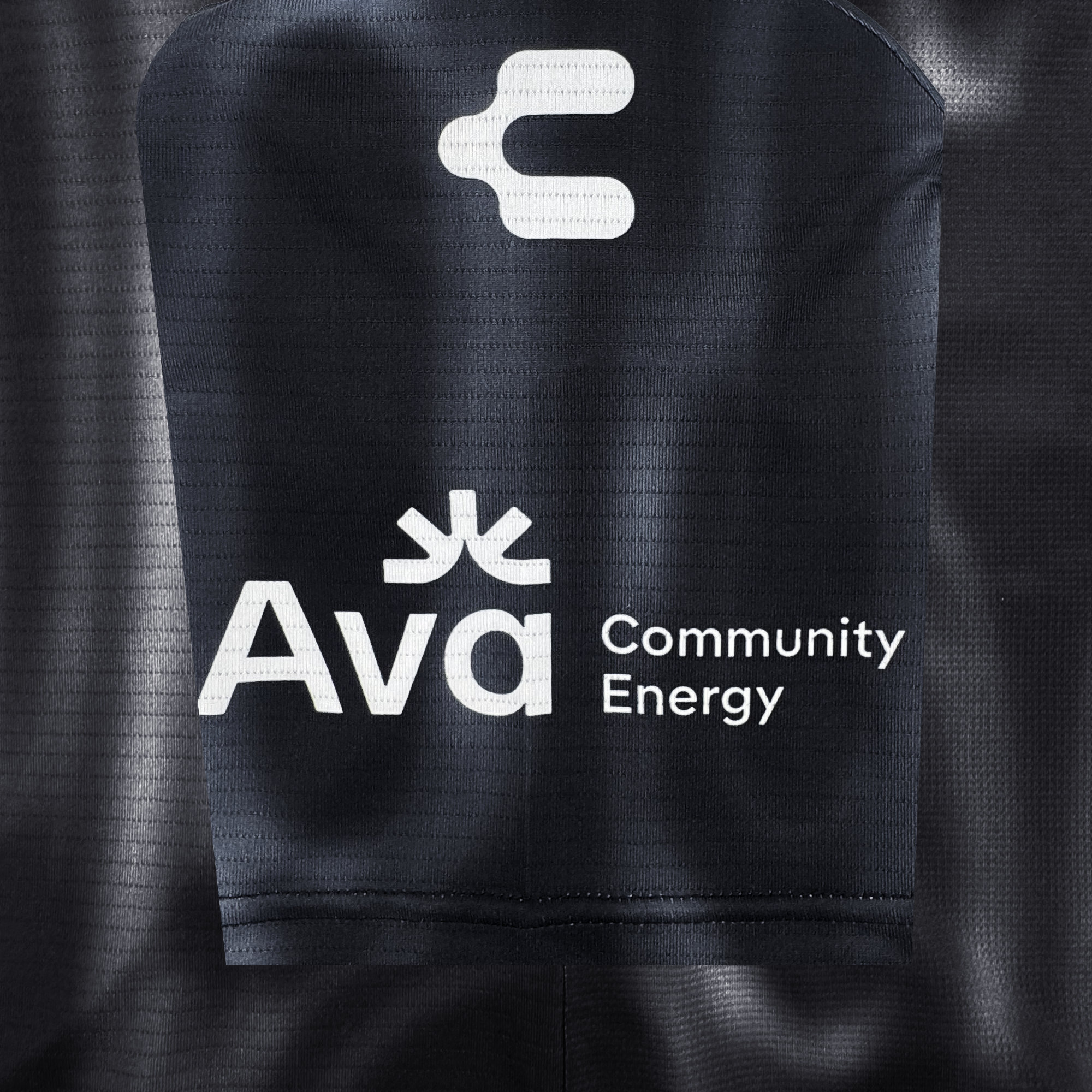 Close-up of a dark-colored Custom 2026 OAKLAND SOUL SC SPIRIT JERSEY by Oakland Soul, showing white logos and text: a stylized energy symbol above "Ava" with "Community Energy" beside it. The fabric is perforated with visible seams.