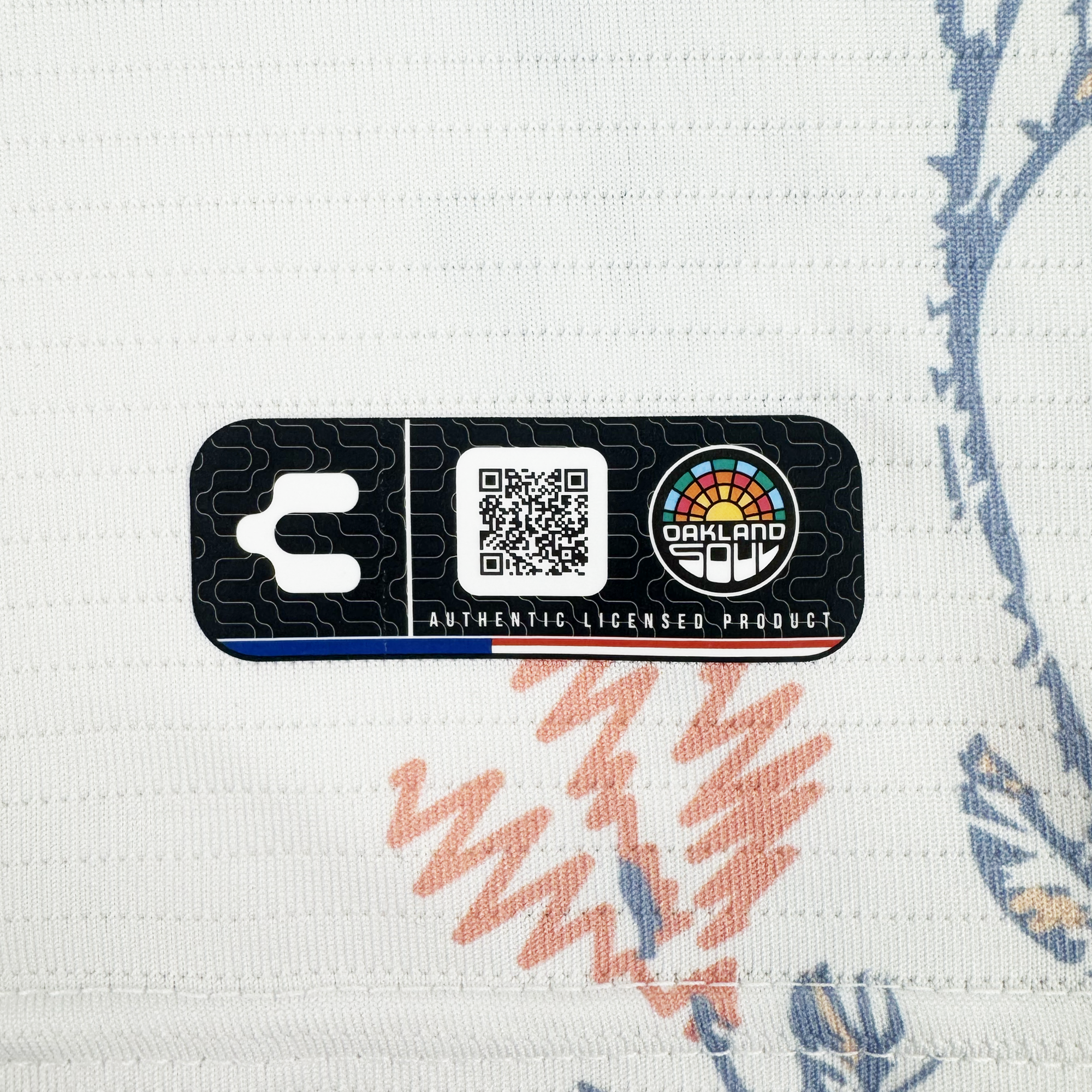 A black rectangular tag with a QR code, the colorful Oakland Soul logo, and “AUTHENTIC LICENSED PRODUCT” text rests on the high-quality polyester 2026 OAKLAND SOUL SC FORM JERSEY by Oakland Soul atop textured white fabric with red and blue abstract patterns.