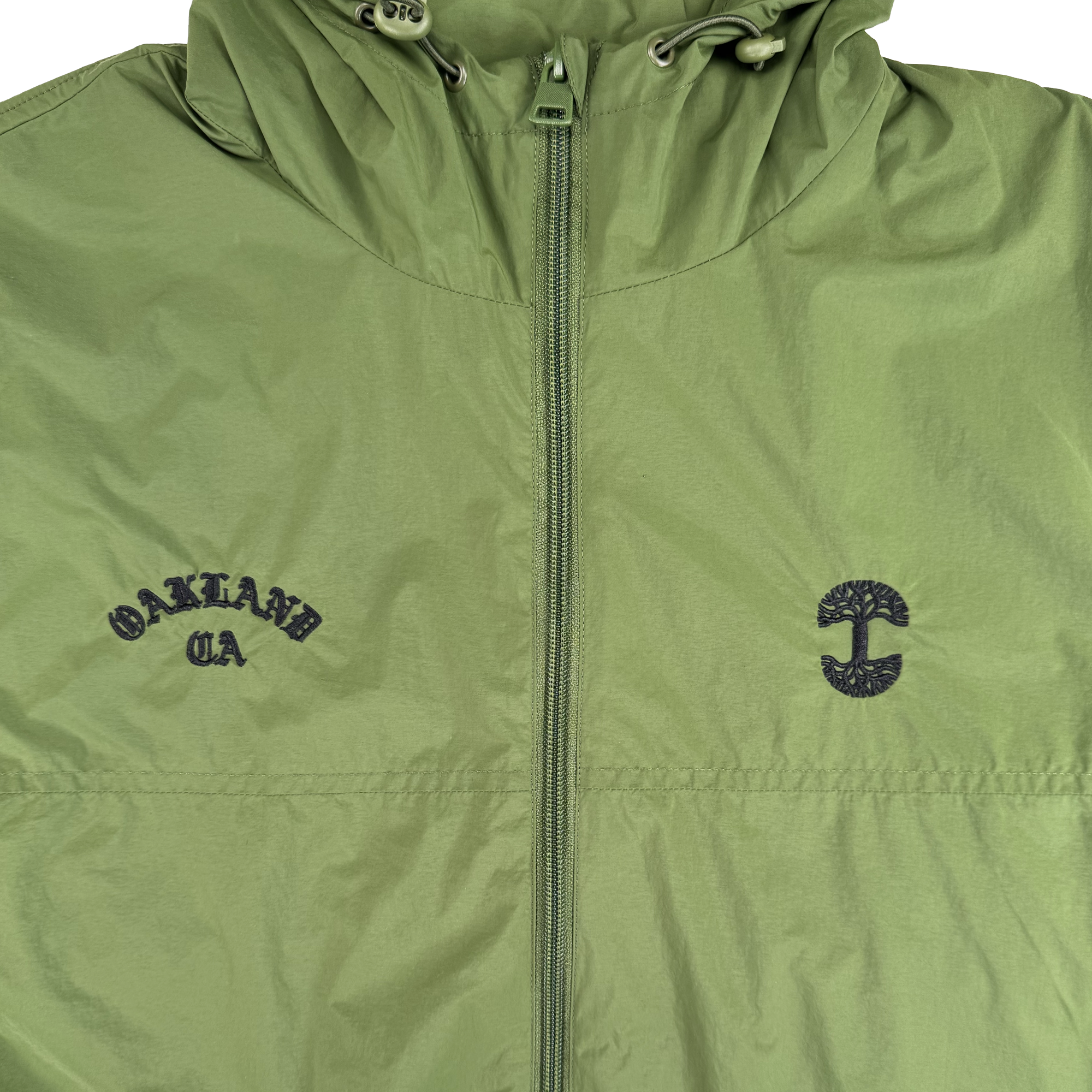 The Susurrus Track Top by Oaklandish is a light green nylon jacket with a classic fit, center zipper, and drawstring hood. "OAKLAND CA" is embroidered in black on the left chest, and a stylized tree appears on the right—an outerwear standout.