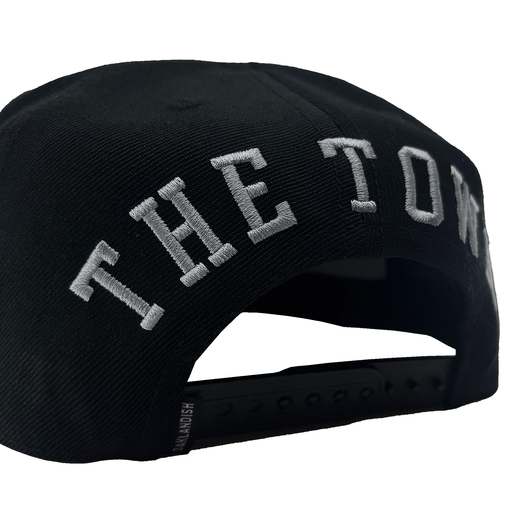 The Town Aft Snapback from Oaklandish is a black cap with an adjustable strap and snap buttons visible at the back. The hat features "THE TOWN" embroidered in large white letters above the strap, along with the iconic Oaklandish tree logo. The background is transparent.