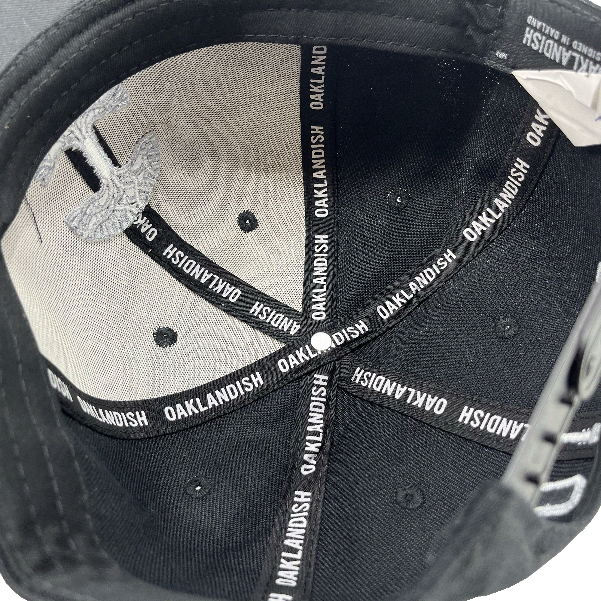The image shows the inside of "The Town Aft Snapback," a black hat from Oaklandish, with the brand name "OAKLANDISH" repeatedly printed on the interior seam taping. The under-visor features a textured and patterned design in light gray, including a partial embroidered Oaklandish tree logo. The overall material appears to be fabric.