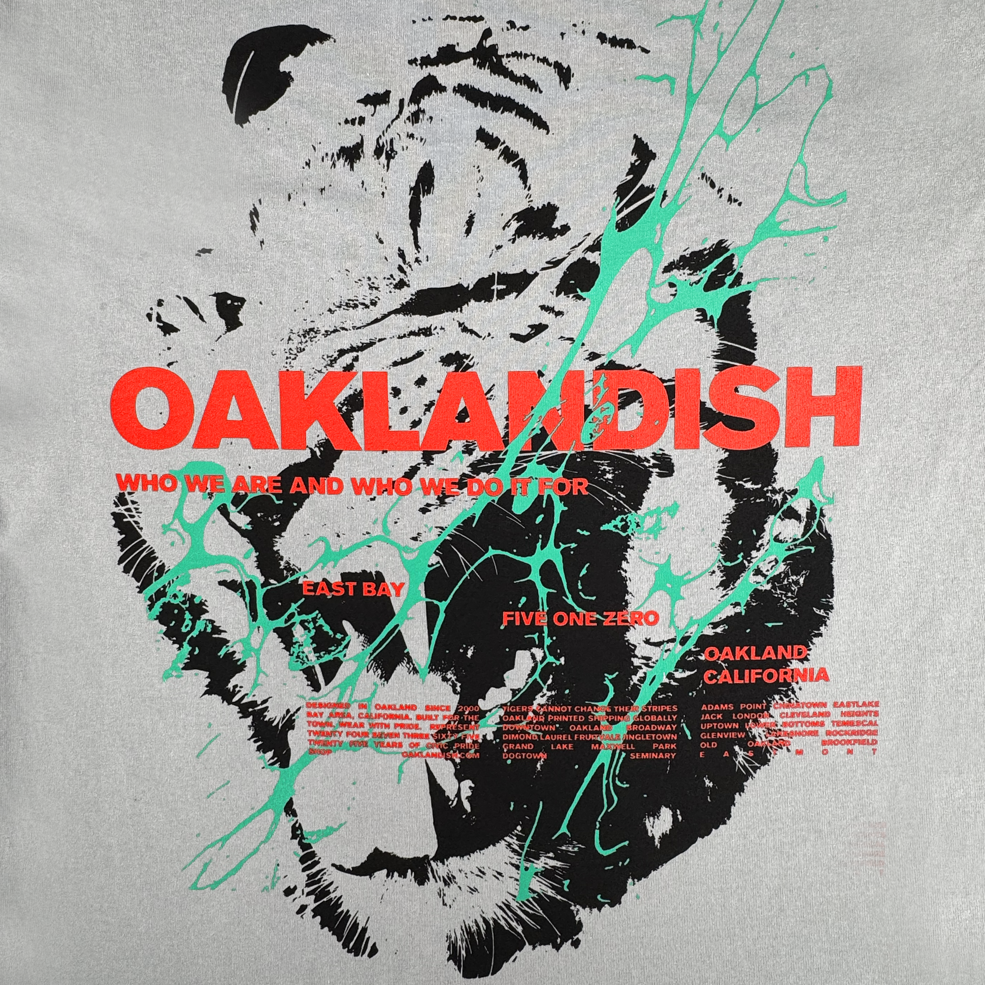 The Tiger Blood Tee by Oaklandish is a vintage grey heavyweight cotton shirt with a black-and-white tiger face, teal paint splatters, bold red "OAKLANDISH," and phrases honoring East Bay, Five One Zero, and Oakland, California.