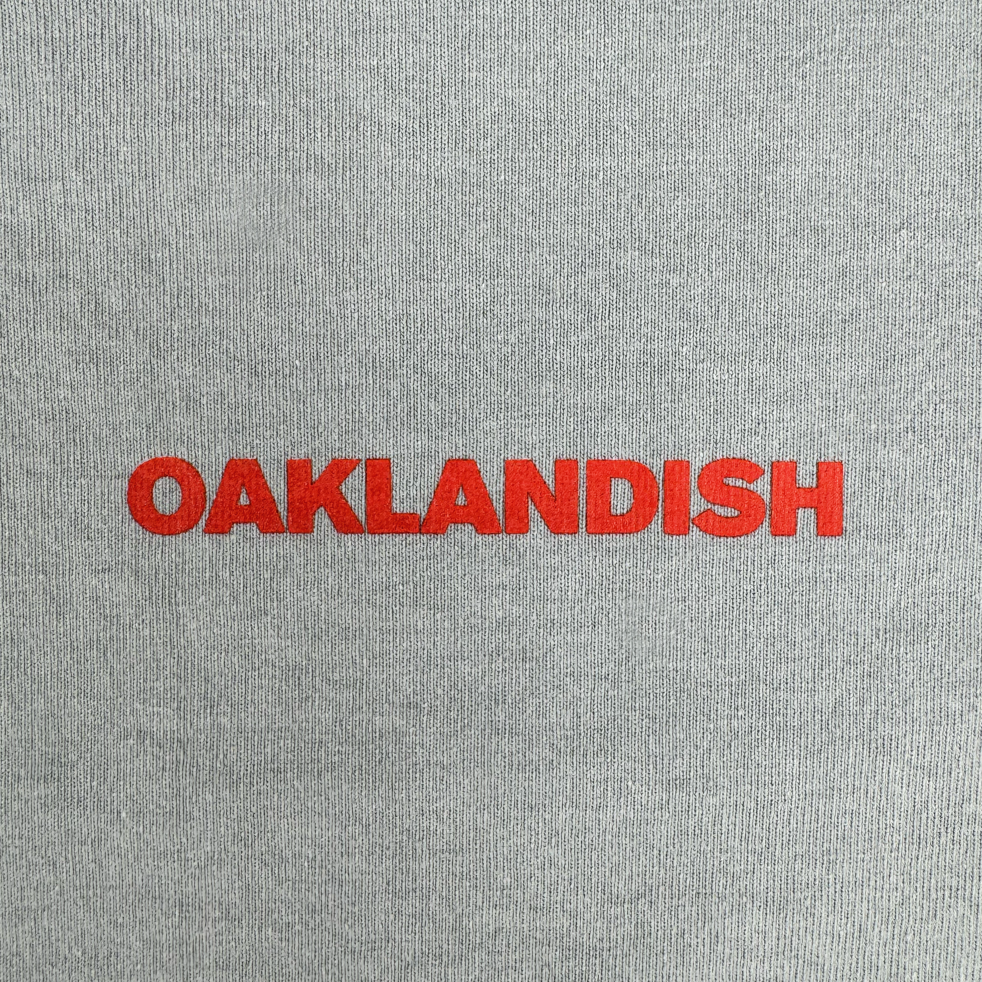 The image features the Oaklandish Tiger Blood Tee: "OAKLANDISH" appears in bold red uppercase letters, centered on a vintage grey, heavyweight cotton shirt with a lightly textured fabric background.