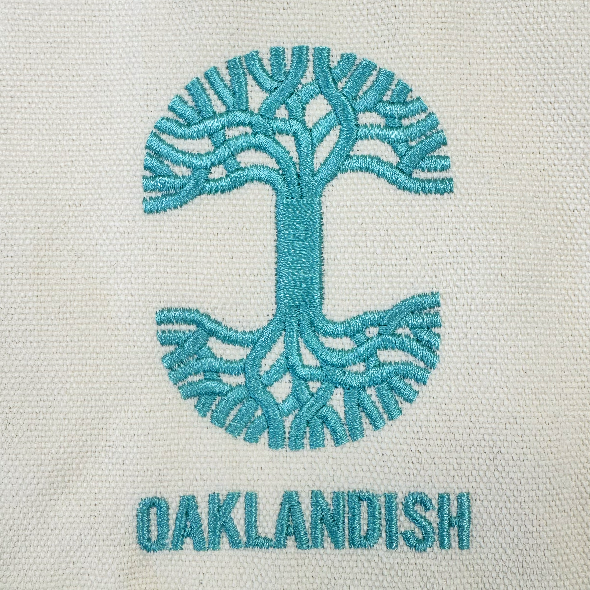Featuring an eye-catching embroidered logo, the Oaklandish Mini Grocery Tote showcases a teal abstract oak tree forming a circular design above the all-caps “OAKLANDISH” brand name, stitched on textured cream fabric for standout style.