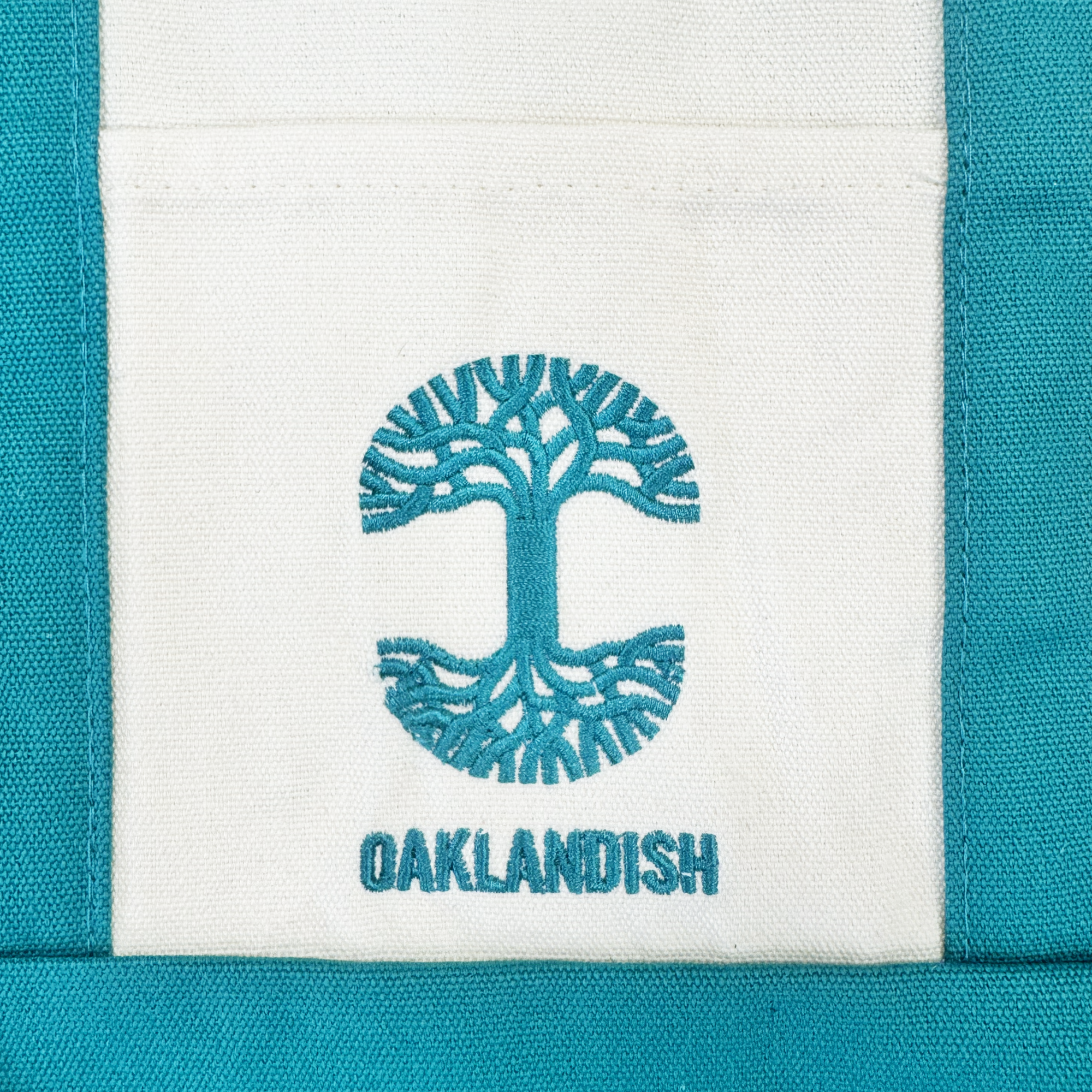 A striking teal blue embroidered tree with expansive roots and branches forms a circle above the word “OAKLANDISH” on a white background, all framed by teal fabric—an eye-catching embroidered logo that makes the Oaklandish Mini Grocery Tote a standout accessory from Oaklandish.