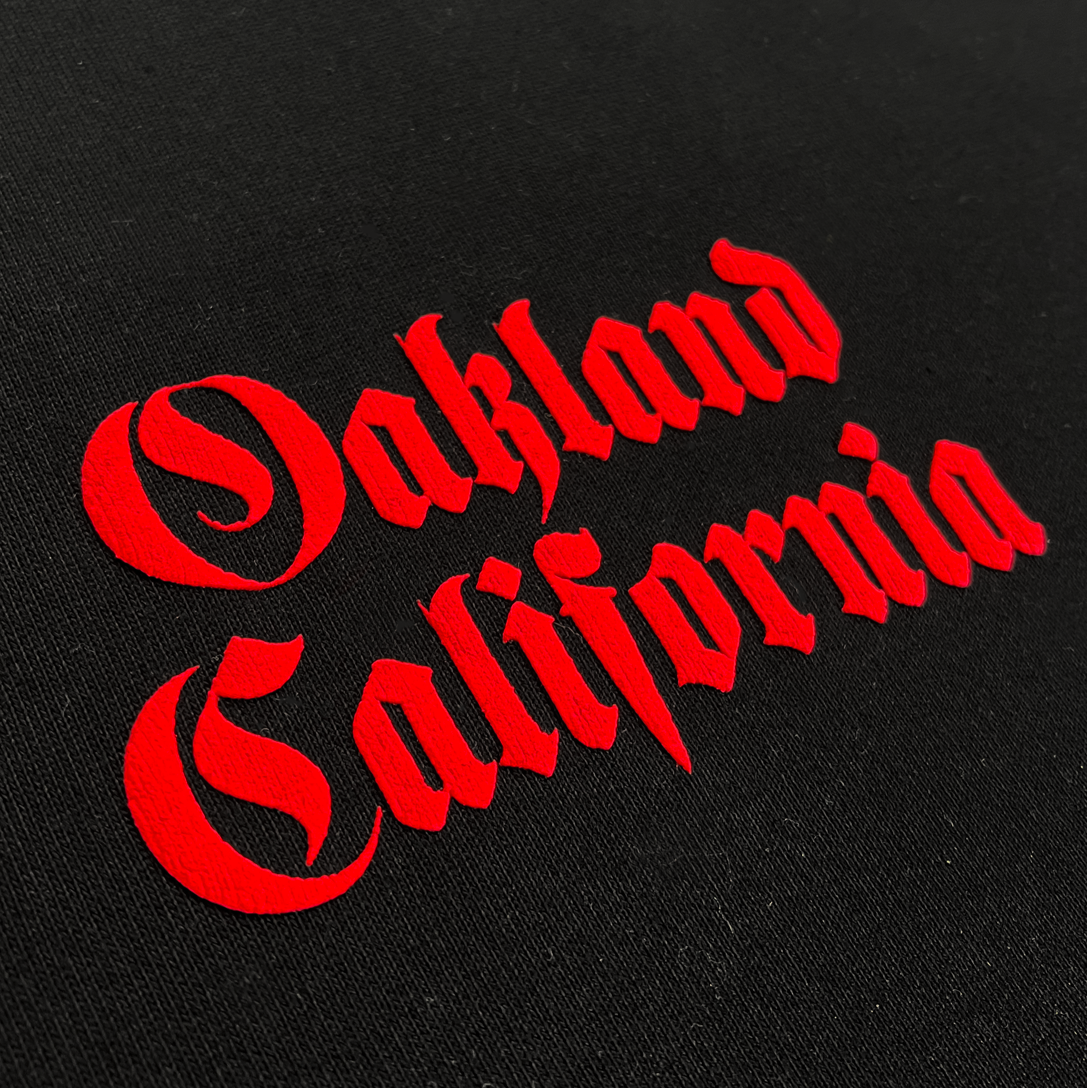 Detailed view of black tee with Oakland California printed centered in red ink.