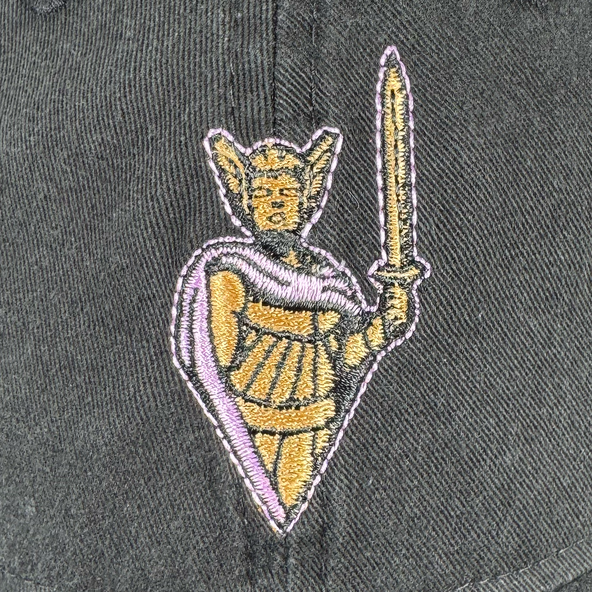 Embroidered patch on dark fabric features a medieval knight in yellow, gold, and purple with a sword and shield—outlined in purple and black. Ideal for the GSV 9TWENTY Dad Hat by GSV, which has an adjustable strap.