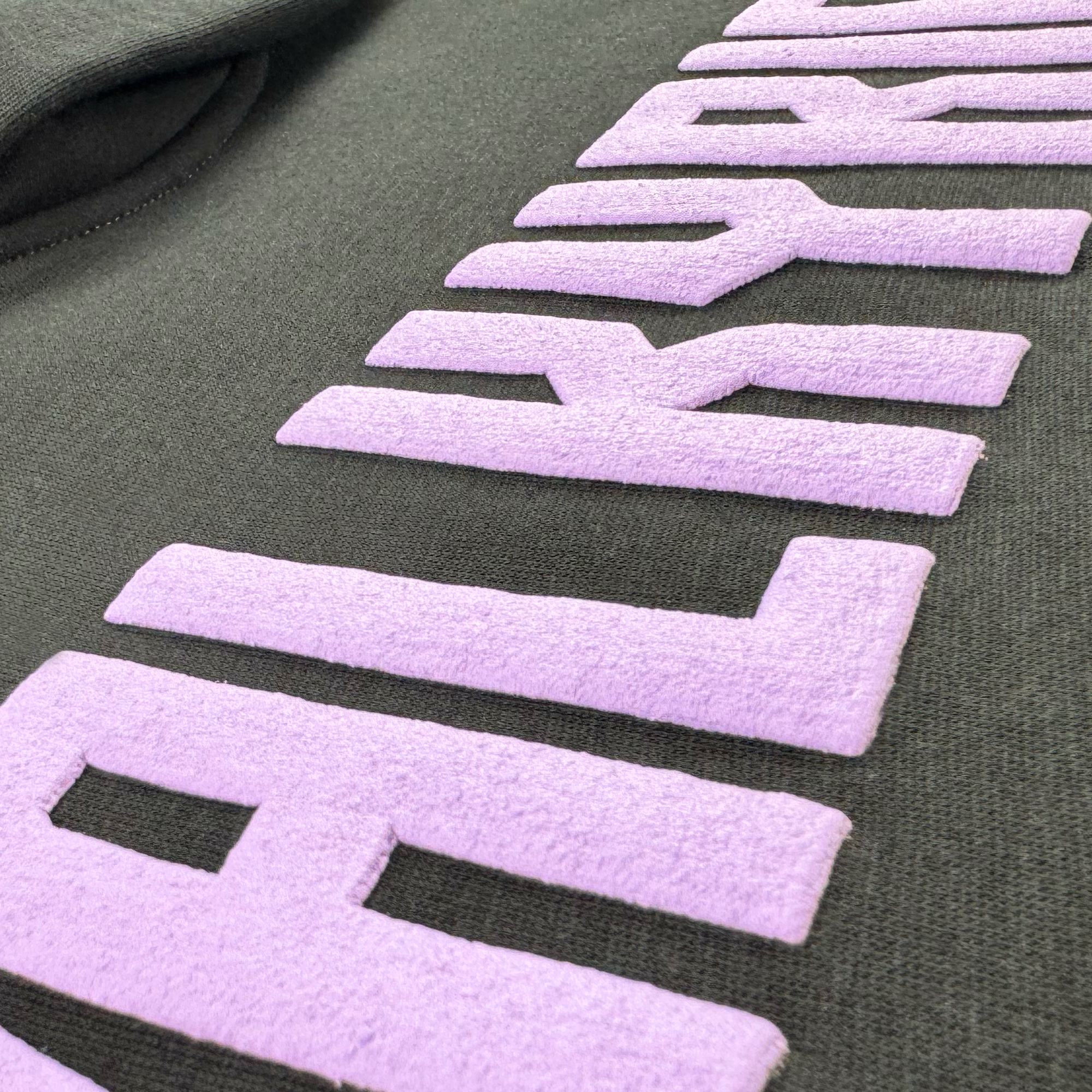Close-up of the Women's Golden State Valkyries Violet Lightning Hoodie by GSV, showing bold, raised light purple "Golden State Valkyries" letters with a velvety texture contrasting against the smooth black fabric. Some lettering is partially out of frame.