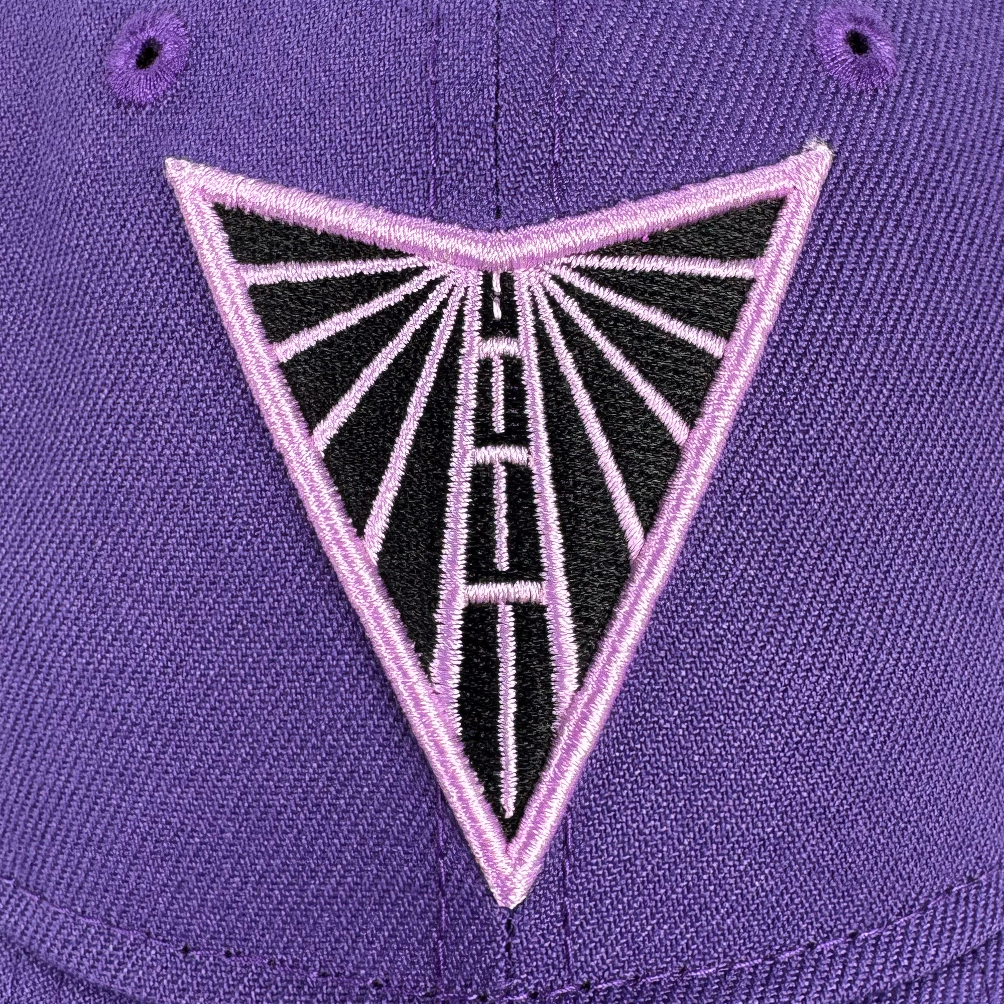 A close-up of the GSV 9TWENTY Dad hat by GSV shows a purple cap with an embroidered triangular patch featuring a pink border, black interior with pink lines, and a central vertical line, resembling a stylized radio tower or sunburst.