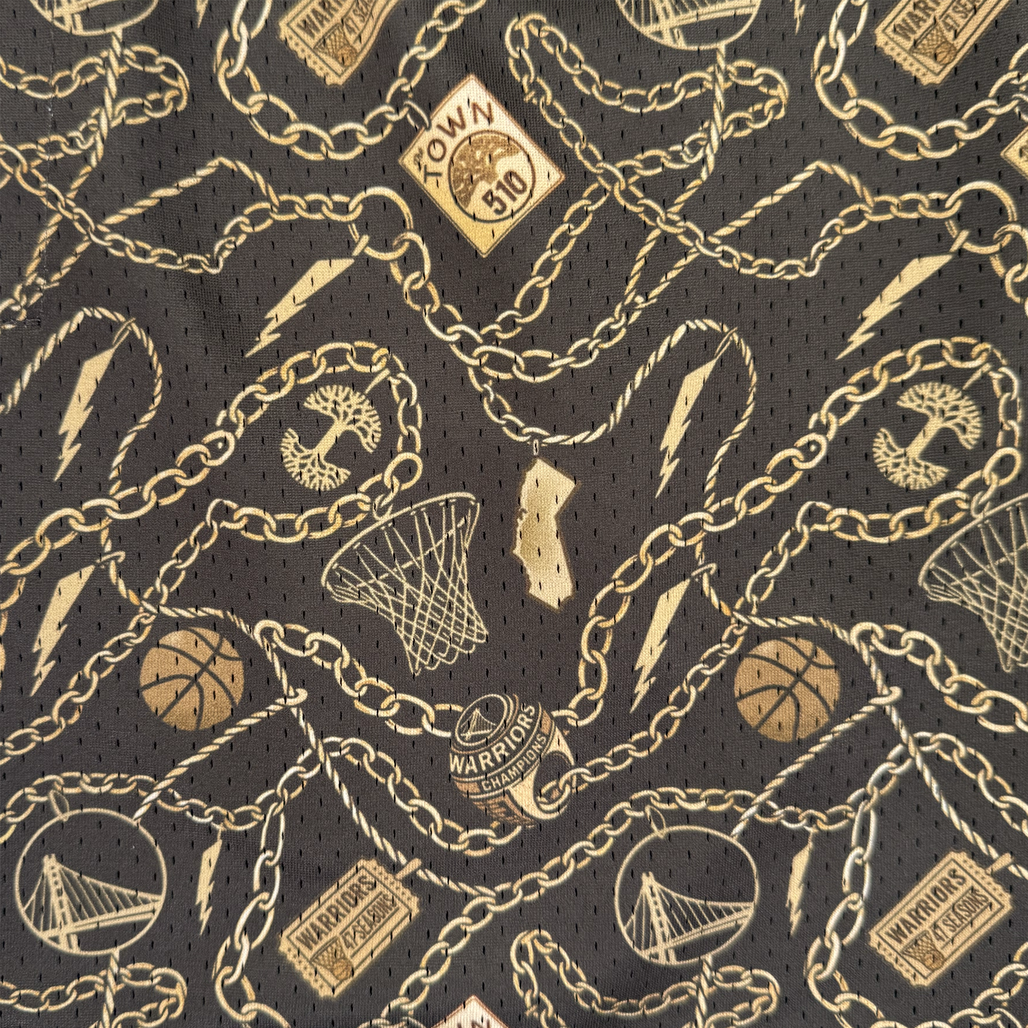 The GSW All Gold Forever Shorts feature a black base with gold chains, basketballs, hoops, and iconic Golden State Warriors elements like "The Town" logo and the Golden Gate Bridge in a repeating Oaklandish-inspired pattern.