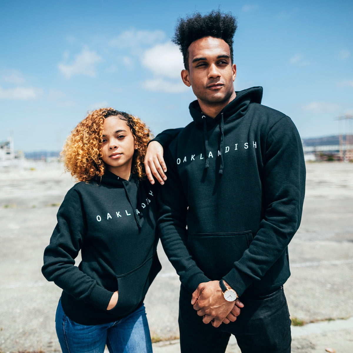 Two people are standing outdoors, both clad in Oaklandish Classic Hoodies with "Oaklandish" printed in white letters on the front. The individual on the left, who has curly hair, is striking a pose with one hand in their pocket. The individual on the right, wearing a men's fit and sporting short hair, stands with their arms crossed. A clear blue sky serves as the backdrop.