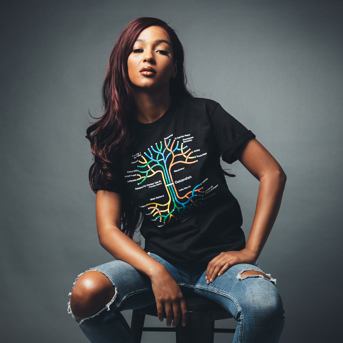 Woman sitting on a stool wearing a black t-shirt with a full-color BART transit map in the shape of the Oaklandish tree logo.