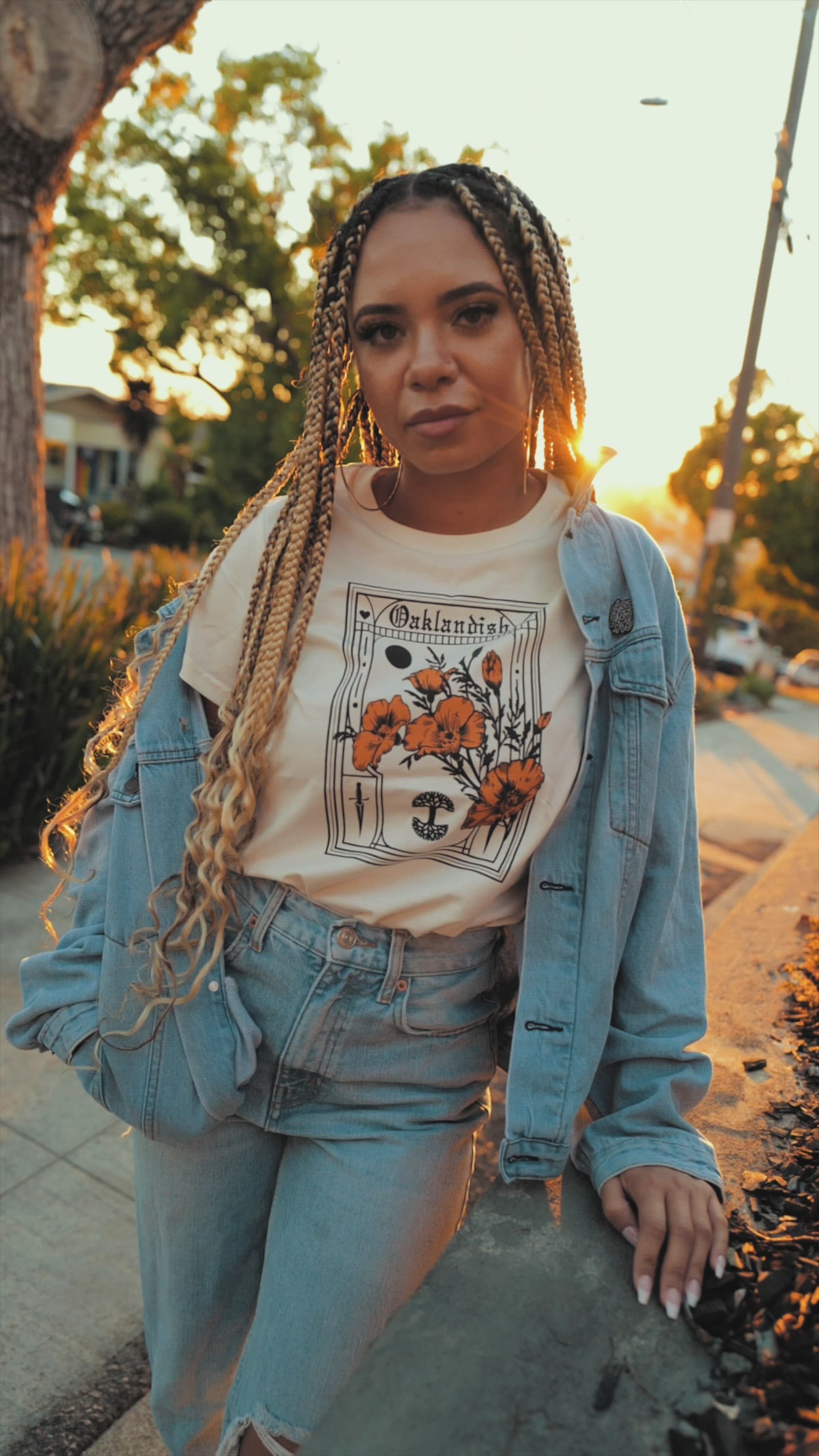 A woman sitting on an Oakland street at sunset leaning against a black car wearing an Oaklandish Blossom t-shirt.